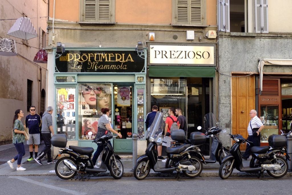 A street scene with old-fashioned stores with moss-green paint in Parma, Italy. Three motorbikes are parked in front of the stores and pedestrians are walking behind them.