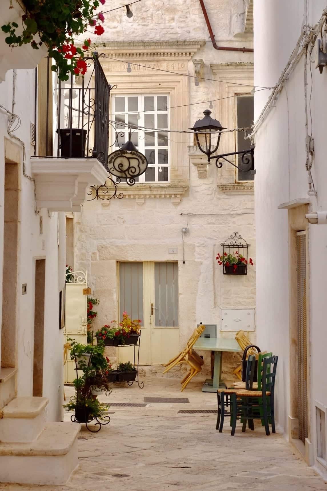 A street in the white city of Locorotondo in Puglia: all white and green. White city walls, green chairs next to a wall, green plants everywhere, black iron streetlamp. A perfect Italian town.