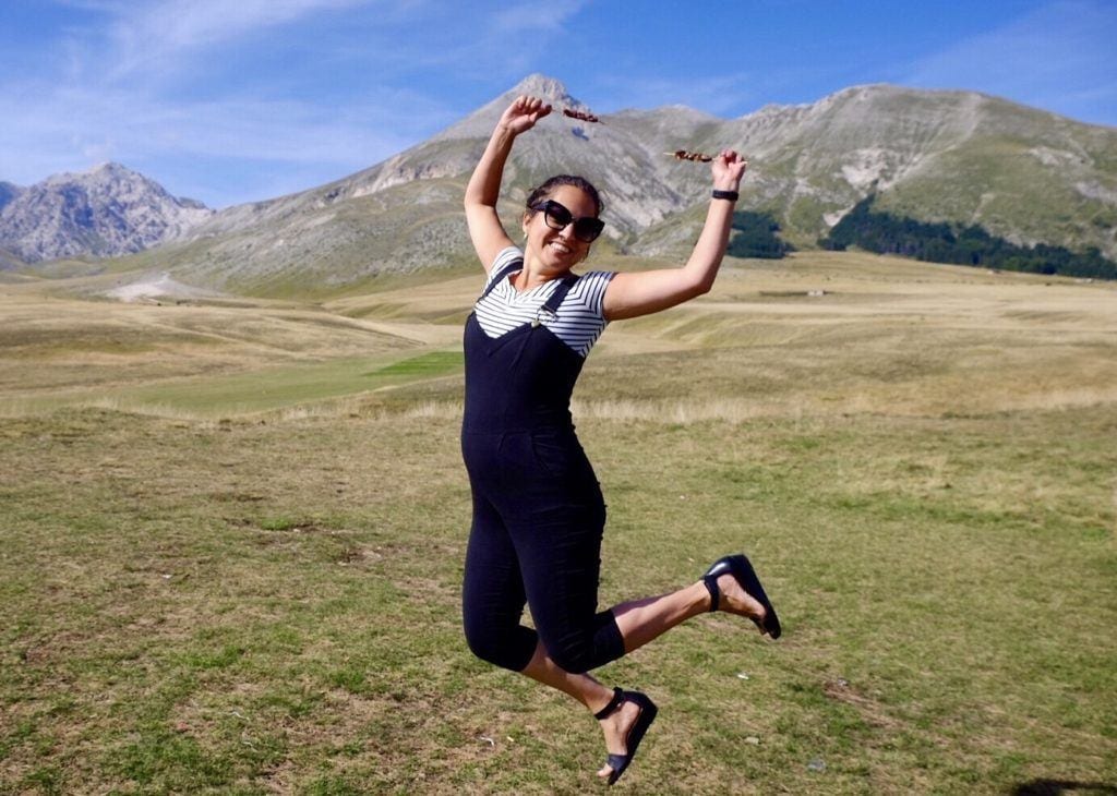 Kate wears overalls and jumps in the air, holding skewers of lamb in each hand, mountains and blue sky behind her, in Gran Sasso National Park.