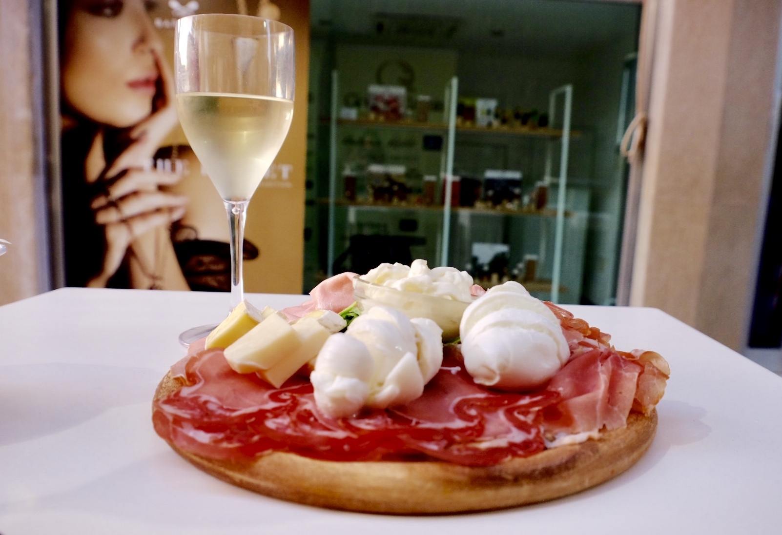 A glass of sparkling white wine next to a tangliere (plate of meat and cheese) in Lecce, Italy: topped with several kinds of ham, burrata mozzarella, and other hams and cheeses.