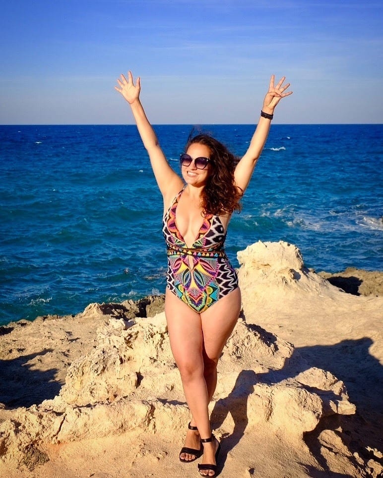 Kate wears a brightly colored and patterned one-piece bathing suit and throws her hands in the air in joy. She's standing on a rocky formation with the bright blue ocean behind her. In Monopoli, Puglia.