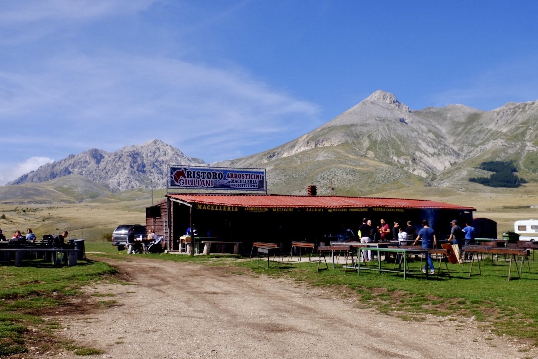 A butcher shop with smoking grills in front of it, in the middle of nowhere in Abruzzo, Italy. Incredible mountains rise in the background underneath a blue sky.