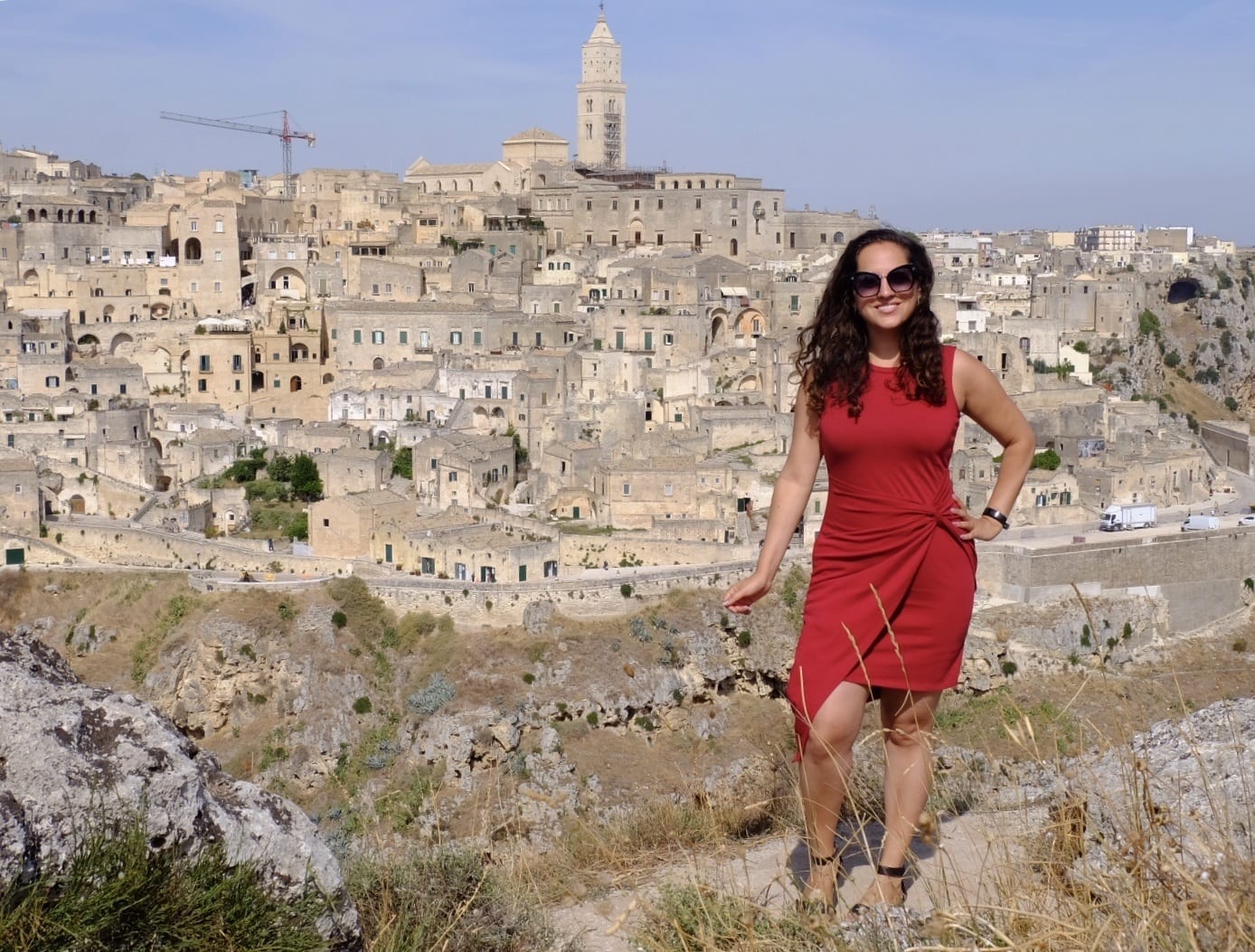 Kate wears a red dress with an asymmetrical hemline and poses in front of the city of Matera: stone towers and homes built on top of a row of sassi (caves).