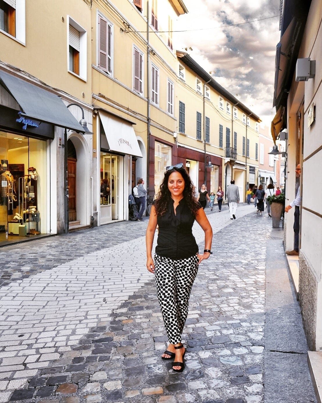 Kate stands on a cobblestone street in Ravenna, Italy, in front of boutique shops in yellow buildings. She wears black and white patterned pants and a sleeveless black blouse.