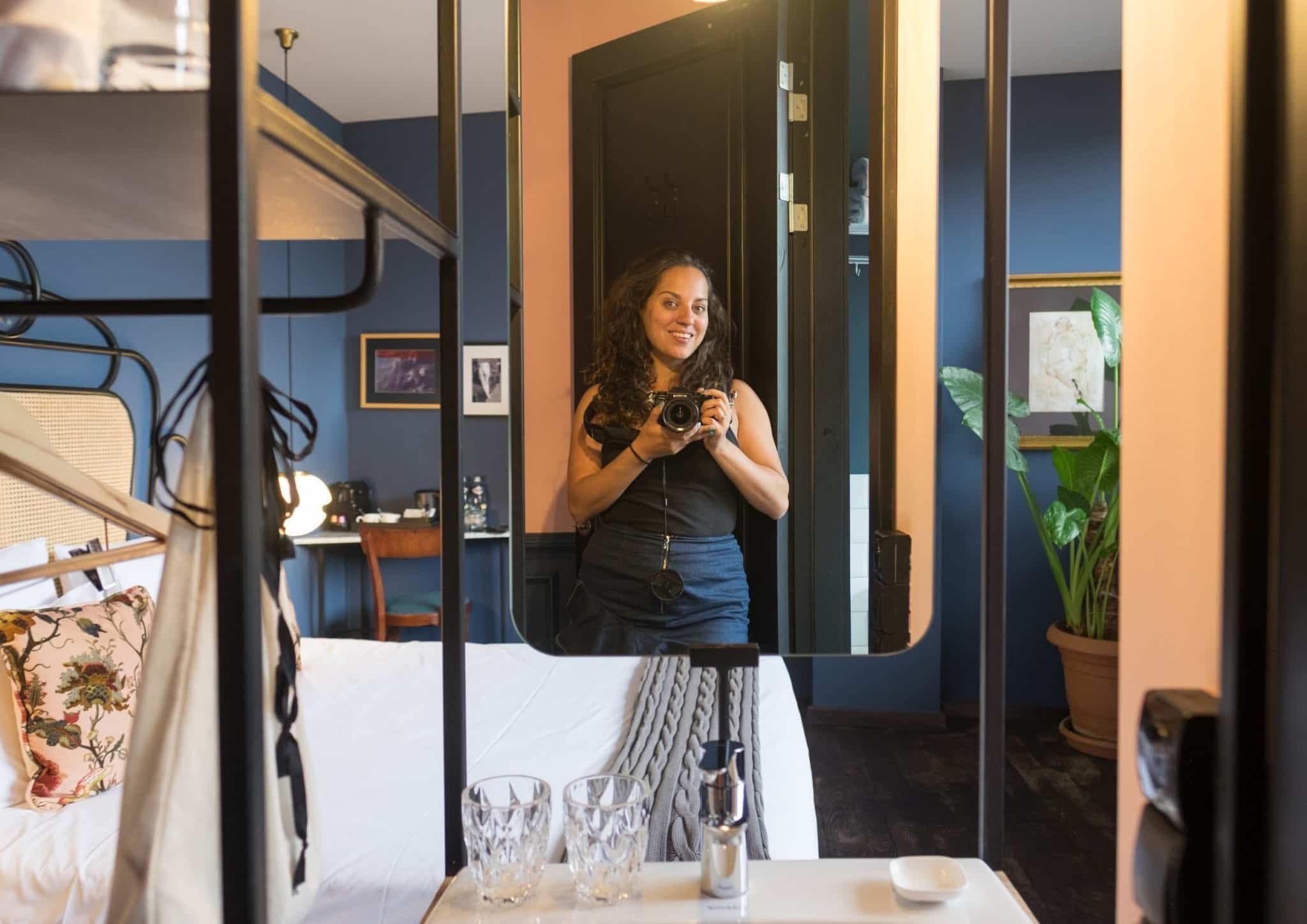 Kate takes a photo in a small mirror above a tiny modern sink; in the background you can see a modern hotel room painted dark blue.