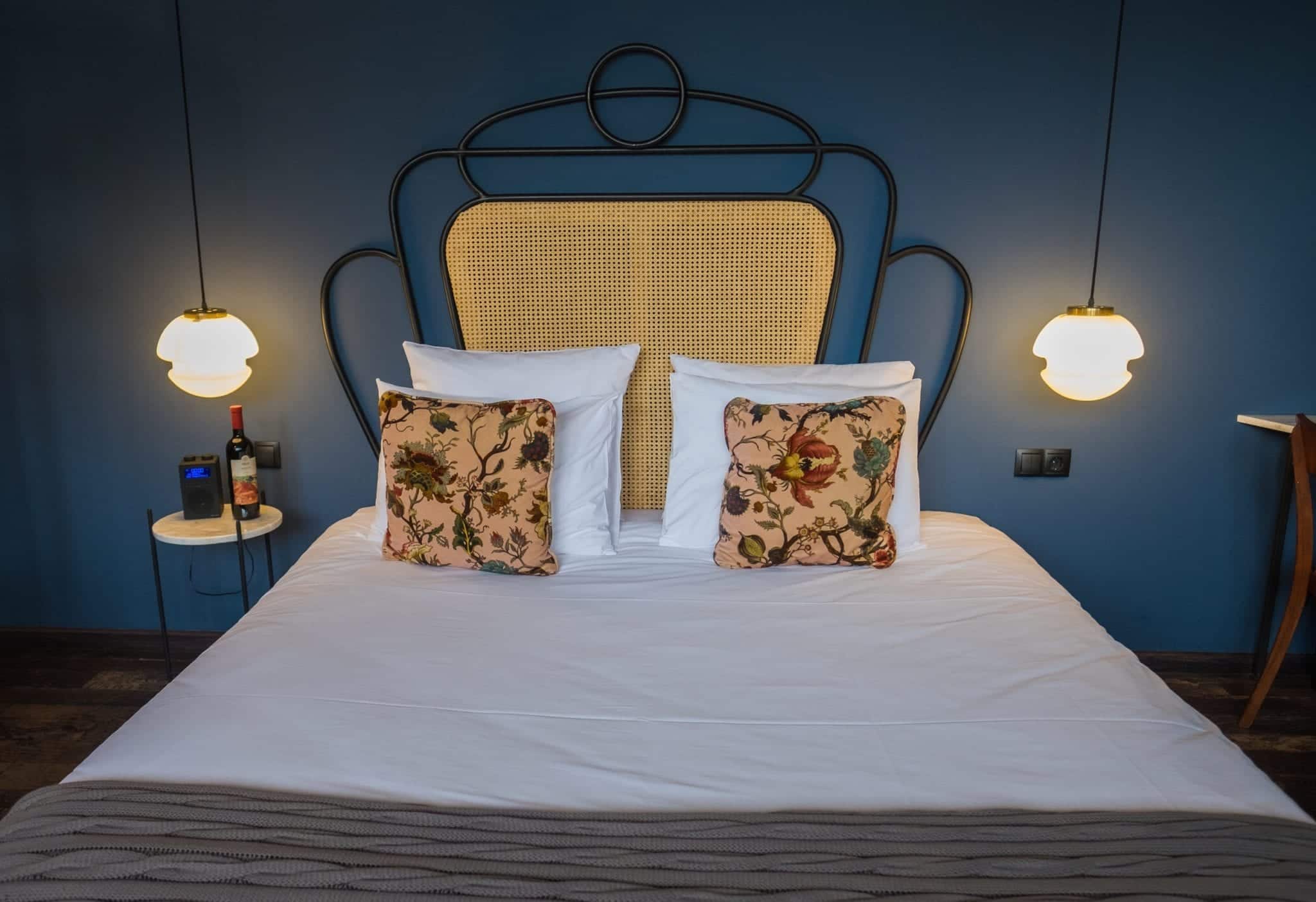 A bed with white sheets, yellow pillows, and a yellow and iron headboard set against a dark blue-green wall with hangings lamps and small modern marble-topped side tables.