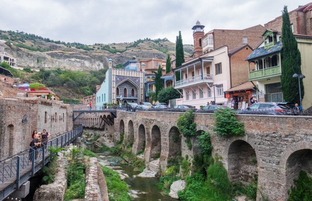 A street scene in Tbilisi's Bath District, with an ancient bridge on one side, people taking photos from a modern iron walkway, and buildings rising up on the hills on each side.