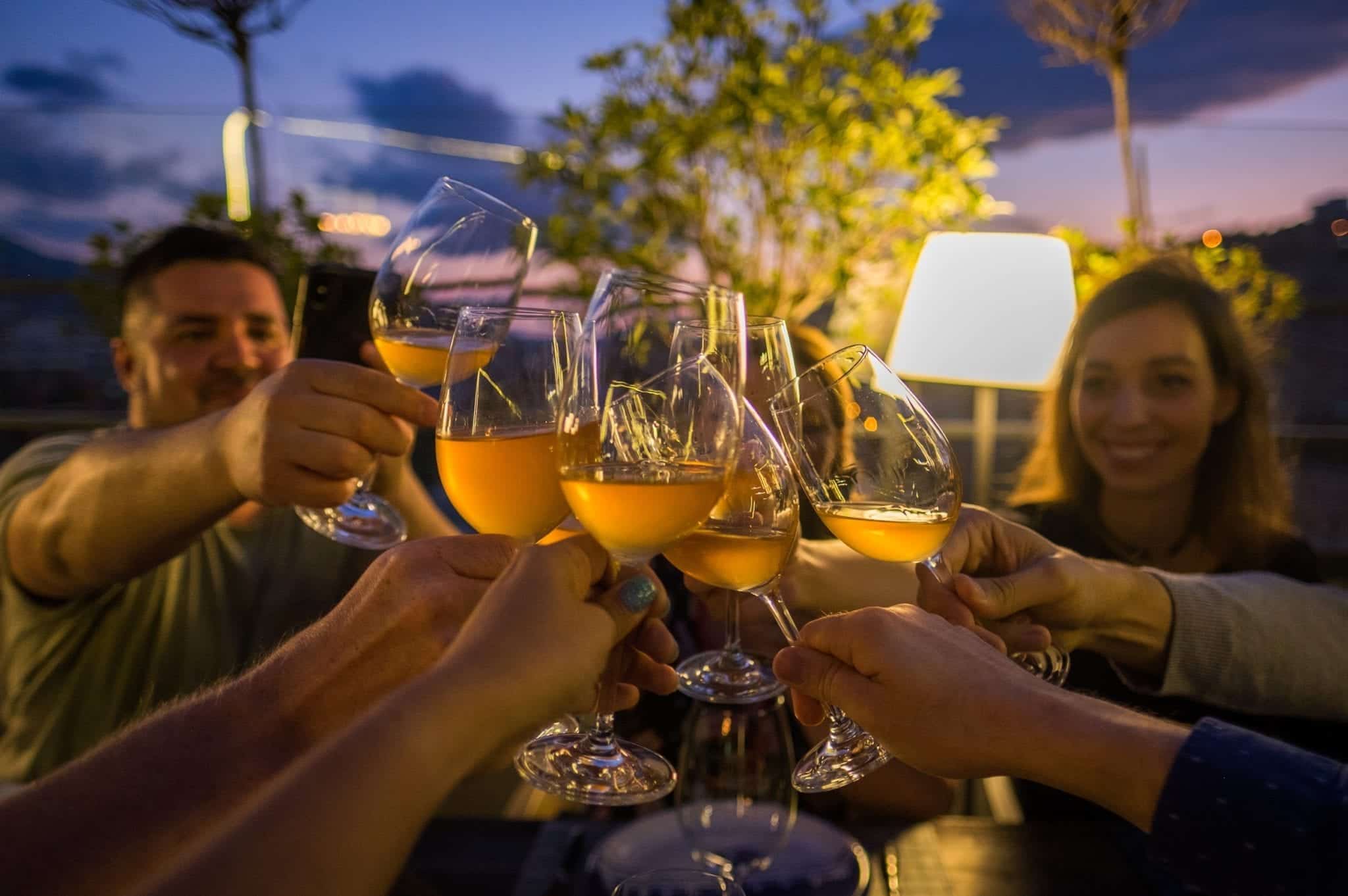 Several hands toasting glasses of orange wine in front of a blue and purple sunset sky.