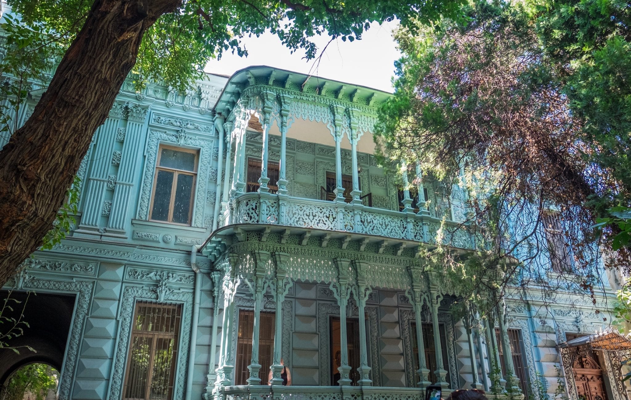 A pale blue house with light sea green trim on the balcony, surrounded by trees.