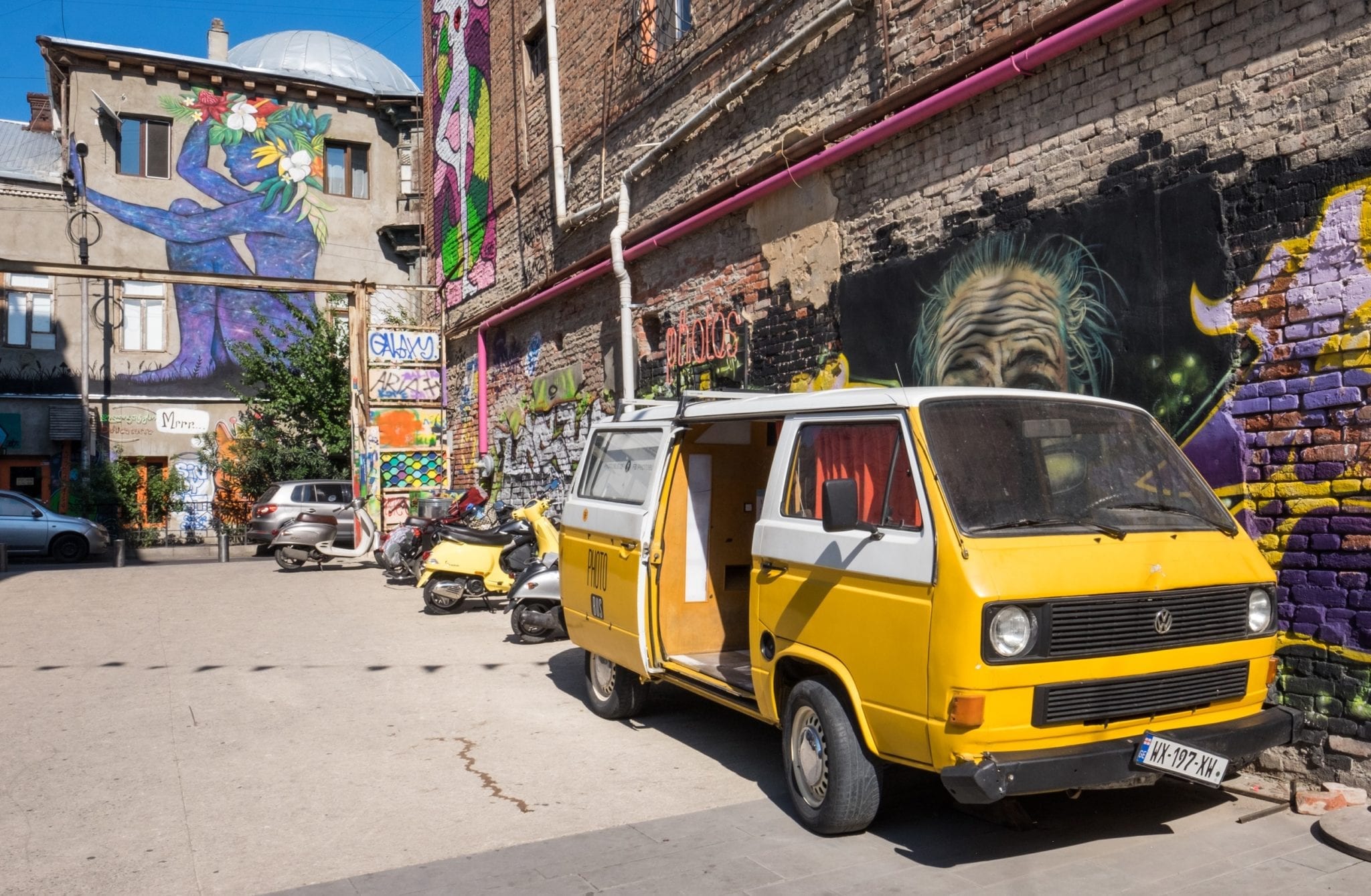 A yellow van parked against a street art-covered wall, with more murals in the background. On the street in Tbilisi.