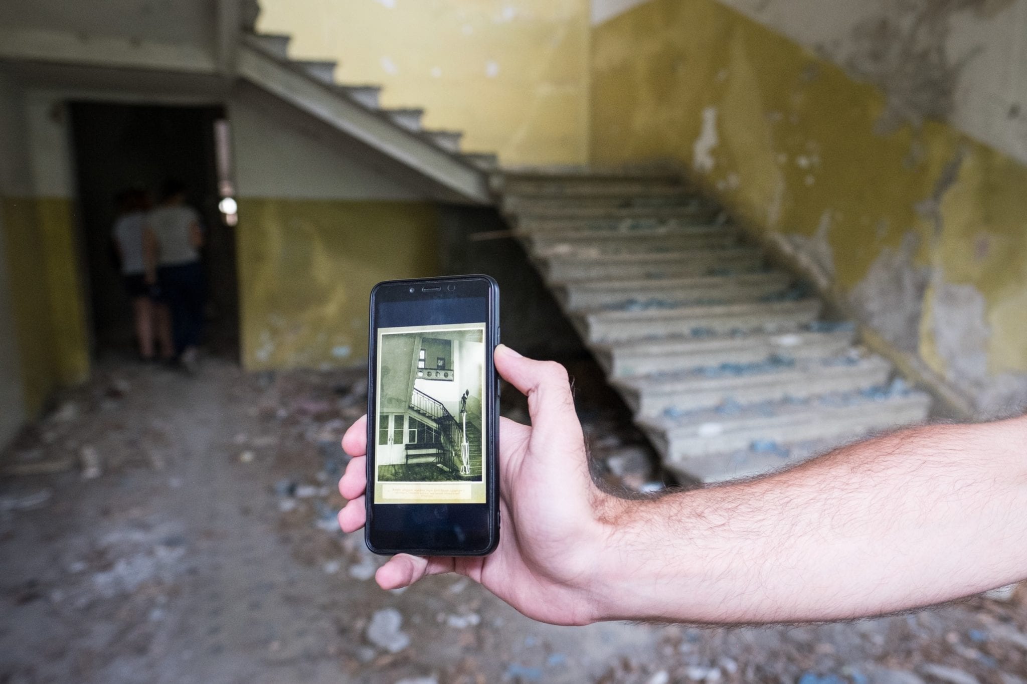 An old, broken down staircase; in the foreground is a hand holding an iPhone that shows the same staircase when it was immaculate.