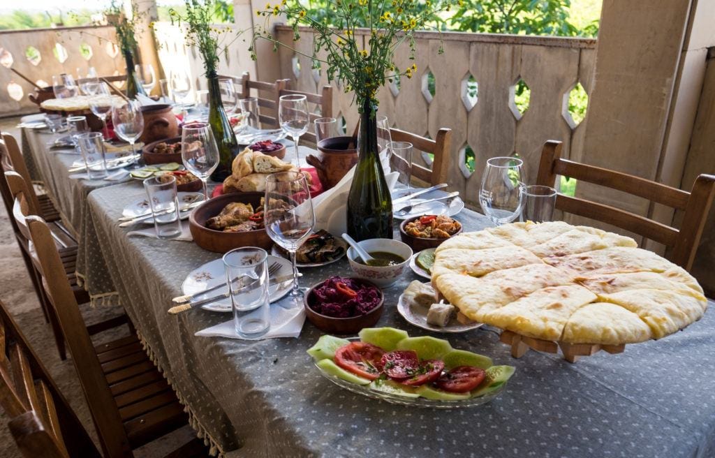 A table covered with plates of Georgian food: khachapuri (cheese pie), tomato walnut cucumber salad, roasted chicken, and lots of wine.
