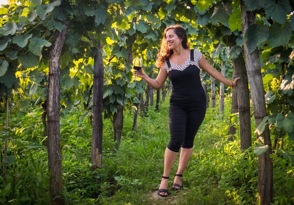 Kate stands in between lines of a vineyard, wearing cropped black overalls and hanging onto a wooden post and smiling as she holds a glass of wine in the other hand.