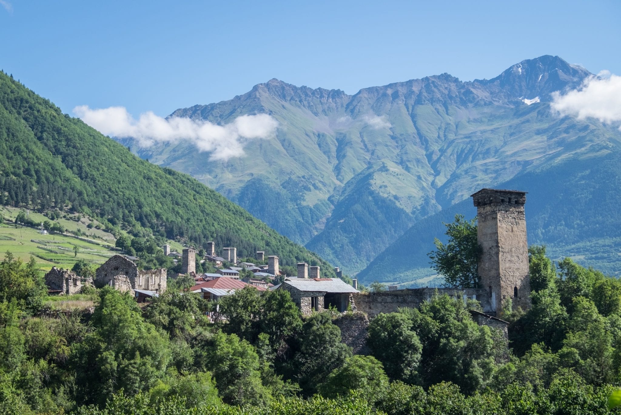 A view over Mestia: mountains in the background, clouds in the sky (lower than the mountains!) and a stone tower on the right and a village with more towers on the left.