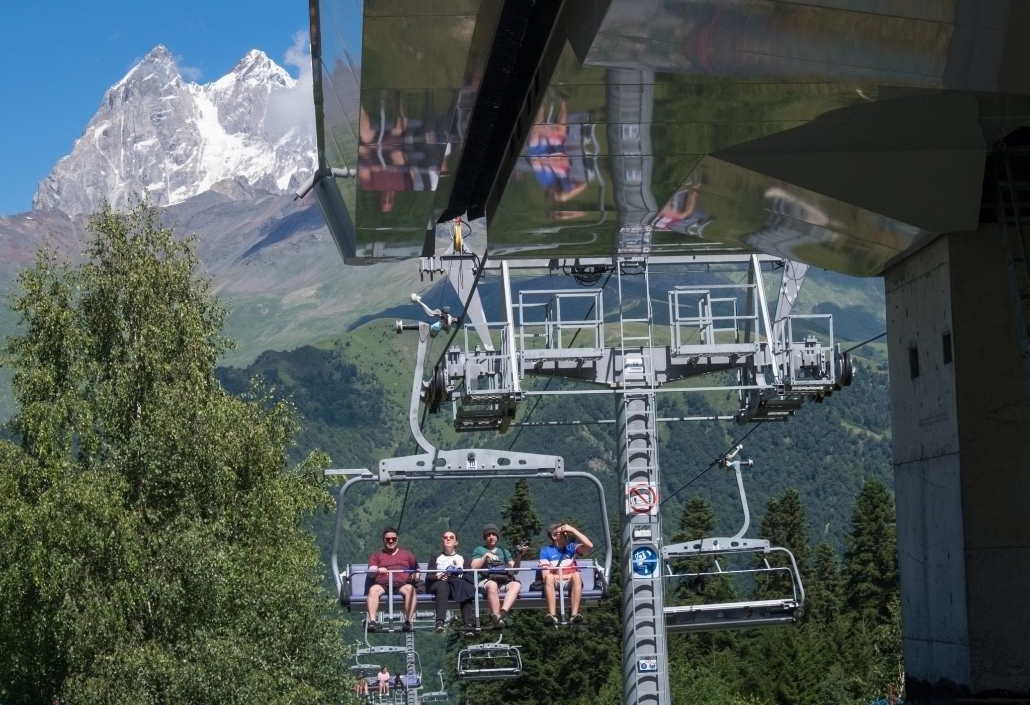 People ride a cable car as a mountain rises up behind them in front of a blue sky.