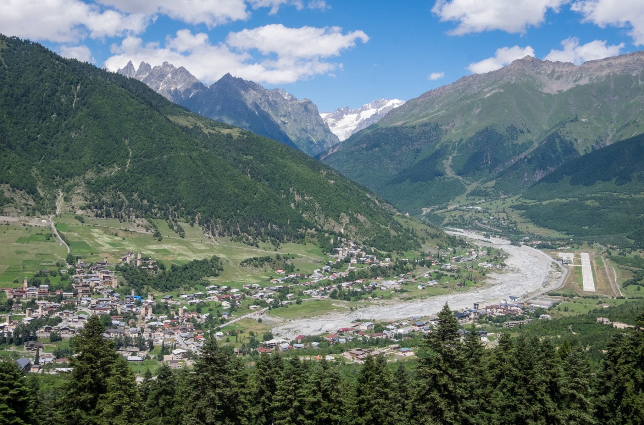A view over the town of Mestia in Svaneti -- huge mountains underneath a blue sky; evergreen trees along the bottom, and a village full of stone towers.