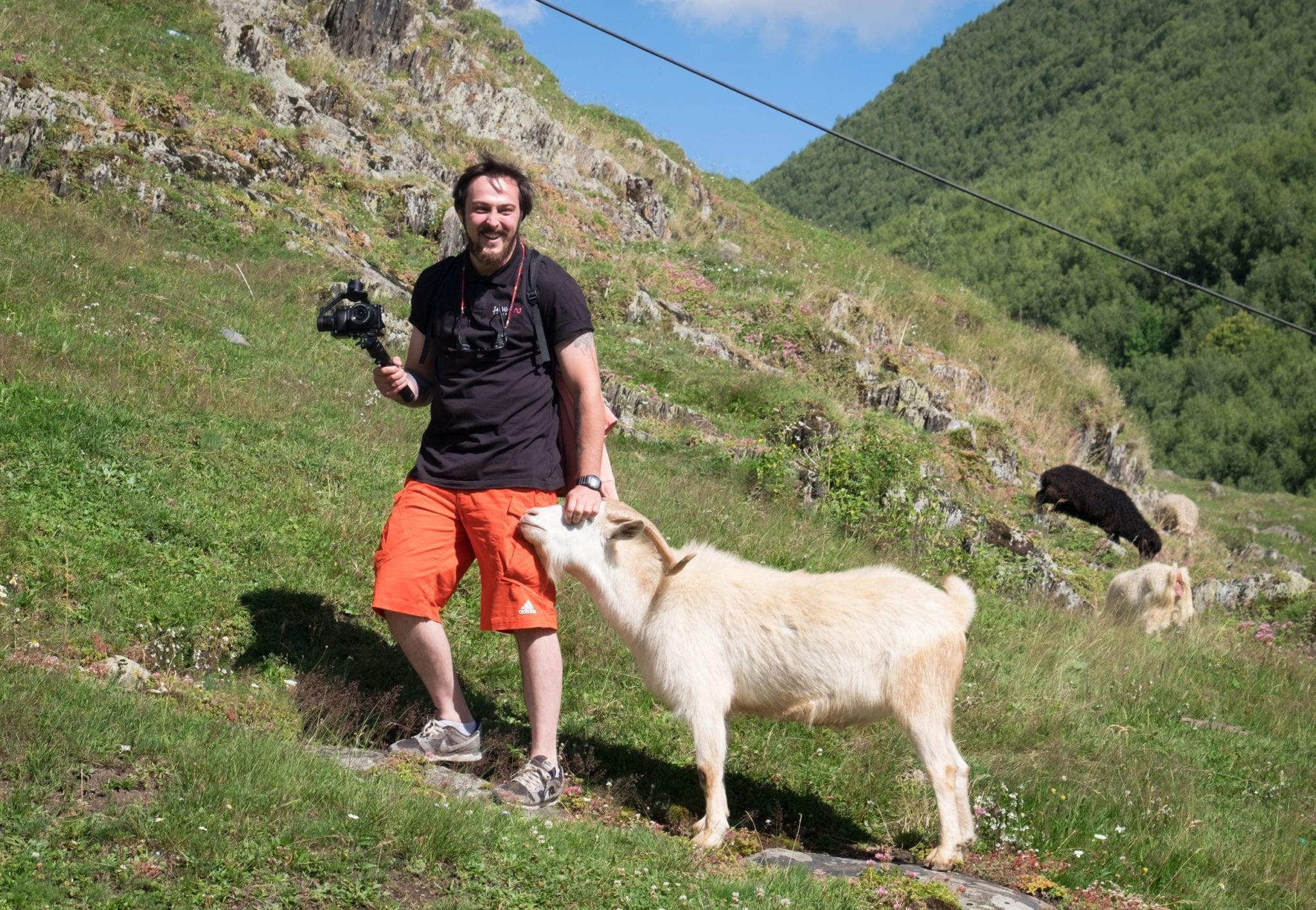 Jayway guide Gio stands on a hillside with a camera and gimbal in one hand and a goat nuzzling his hip. He strokes the goat's head and laughs.