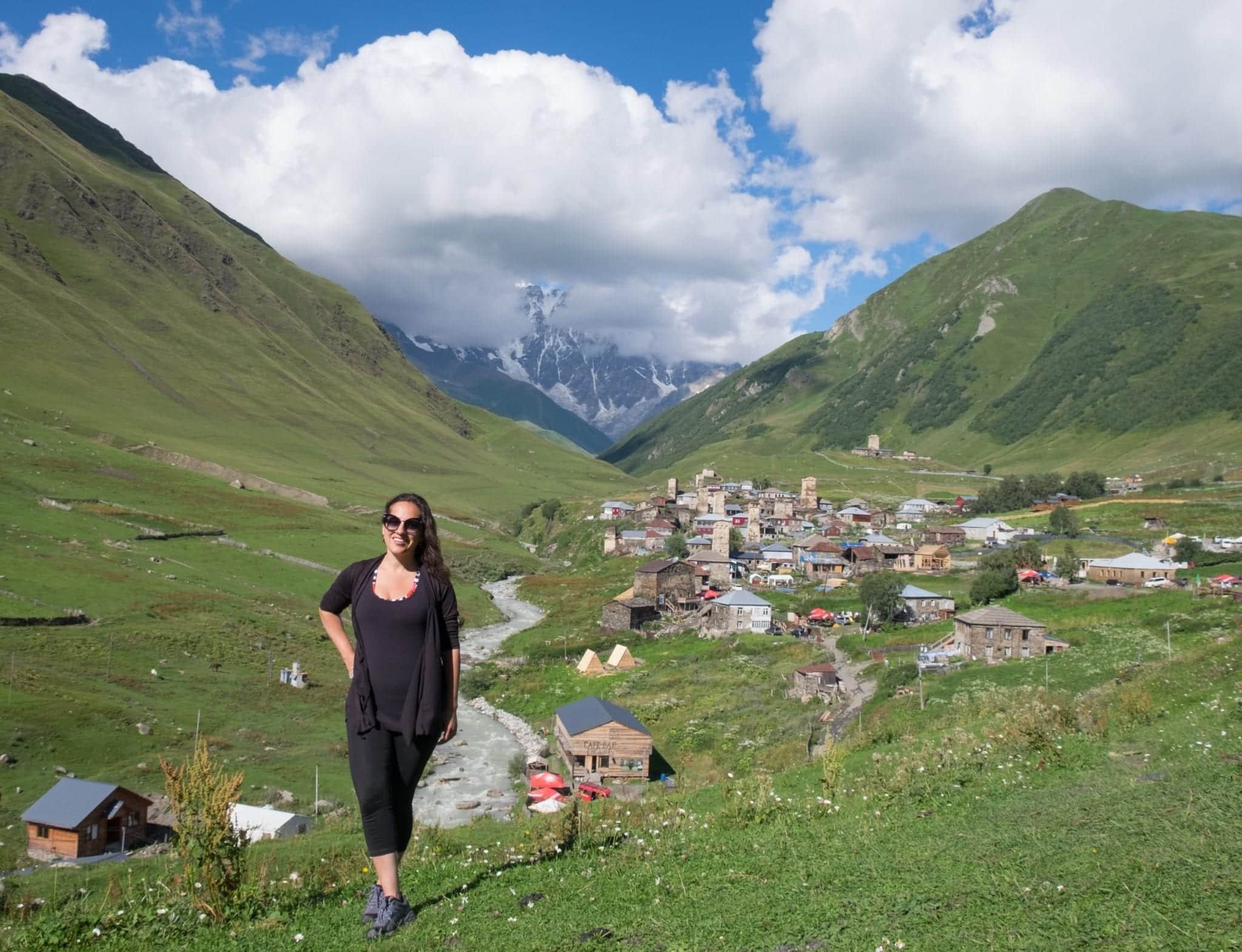 Kate stands on a grassy hill in front of the village of Ushguli in Georgia, with its stone towers. Behind the village is a mountain partially obscured by clouds.