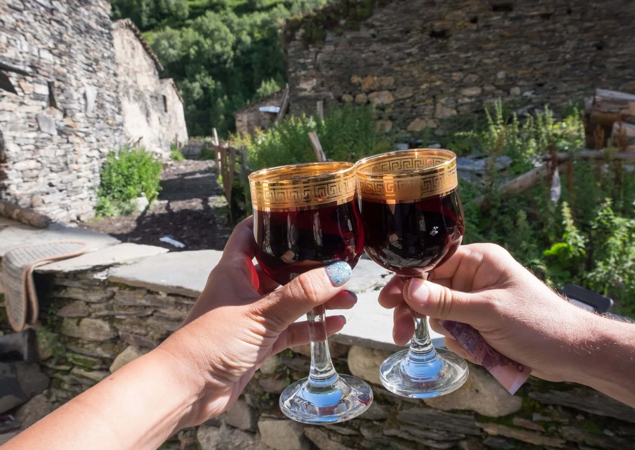 Two hands holding up tiny wine glasses edged with gold, each holding red wine, stone buildings in the distance.