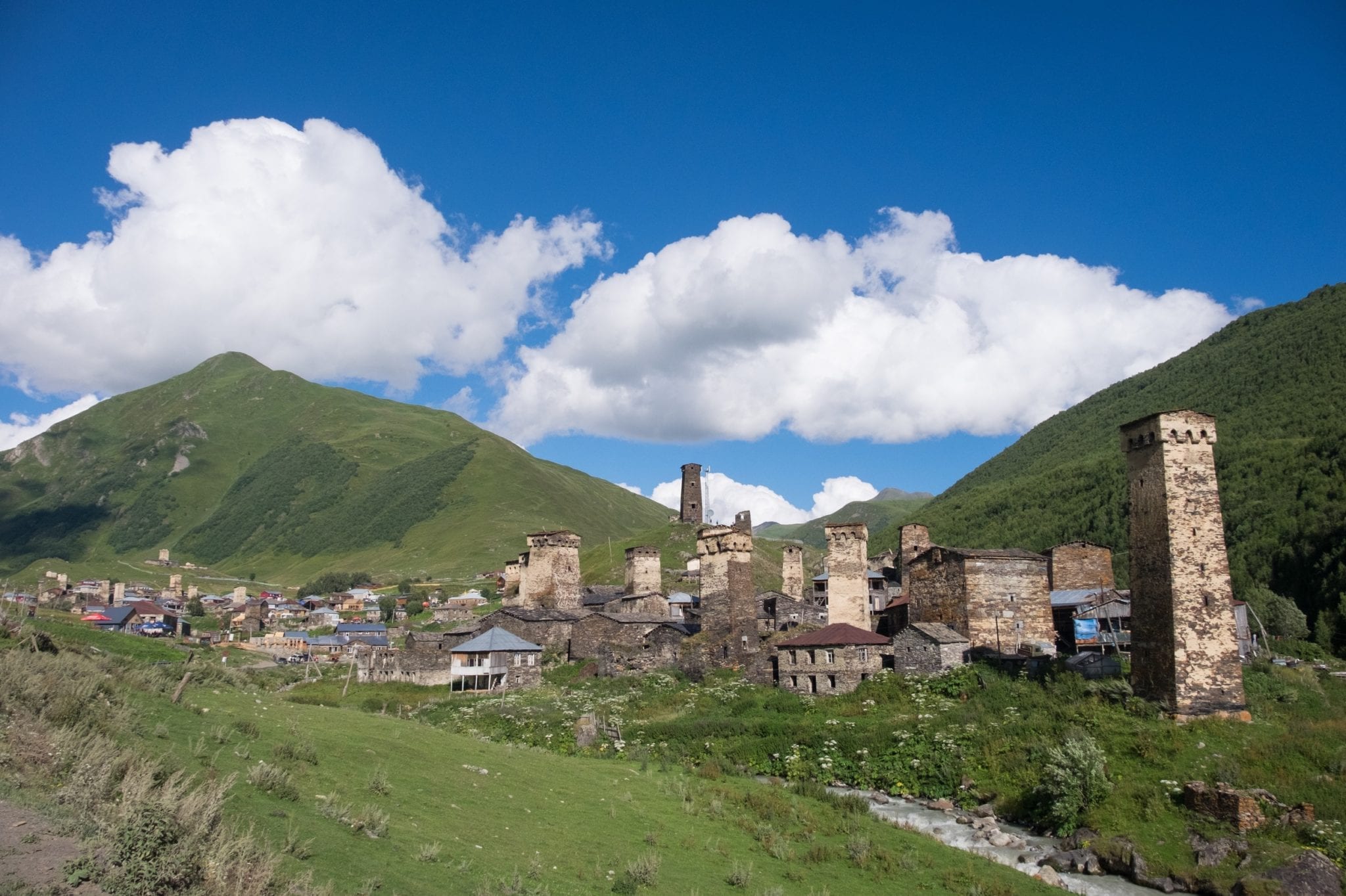 On the right, the village of Chazhashi in Ushguli, stone towers pointing into the air. In the background are green grassy mountains underneath a blue sky with clouds,