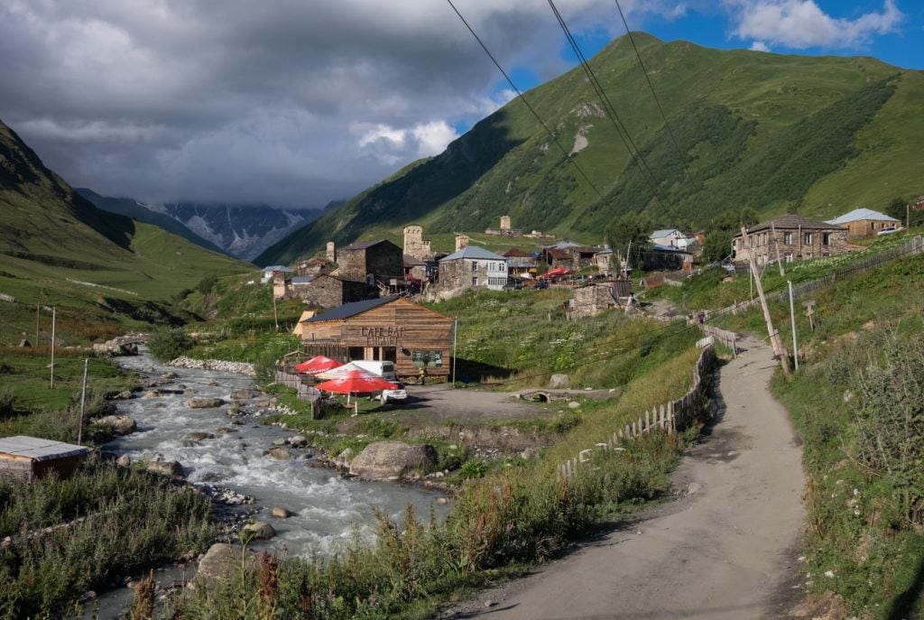 A path leading past a stream in Ushguli, leading to guesthouses and cafes with green mountains in the distance.