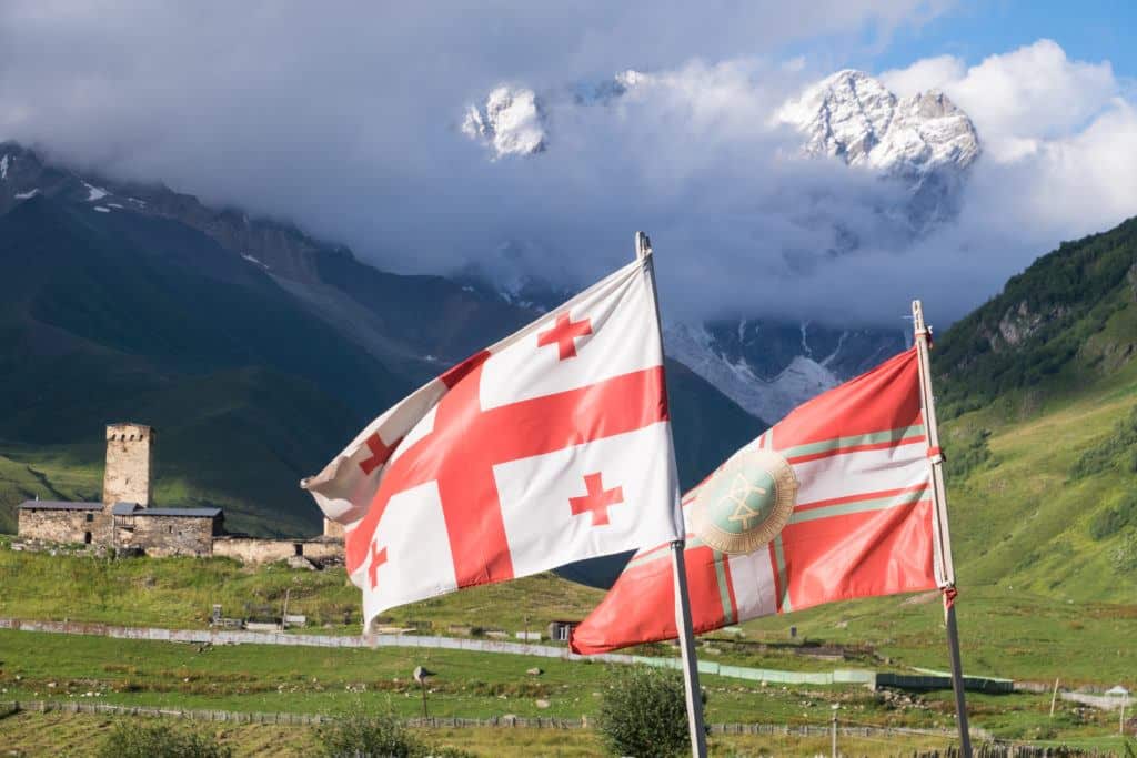The Georgian flag and the Svaneti flag, both red and white, flying side by side with the mountains and stone towers of Ushguli in the background.