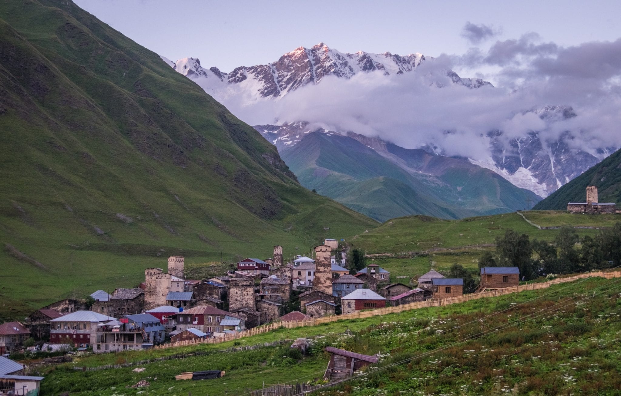 At dusk, the village of Ushguli looking purple in the evening light, surrounded by hills, purple mountains covered by clouds in the background.