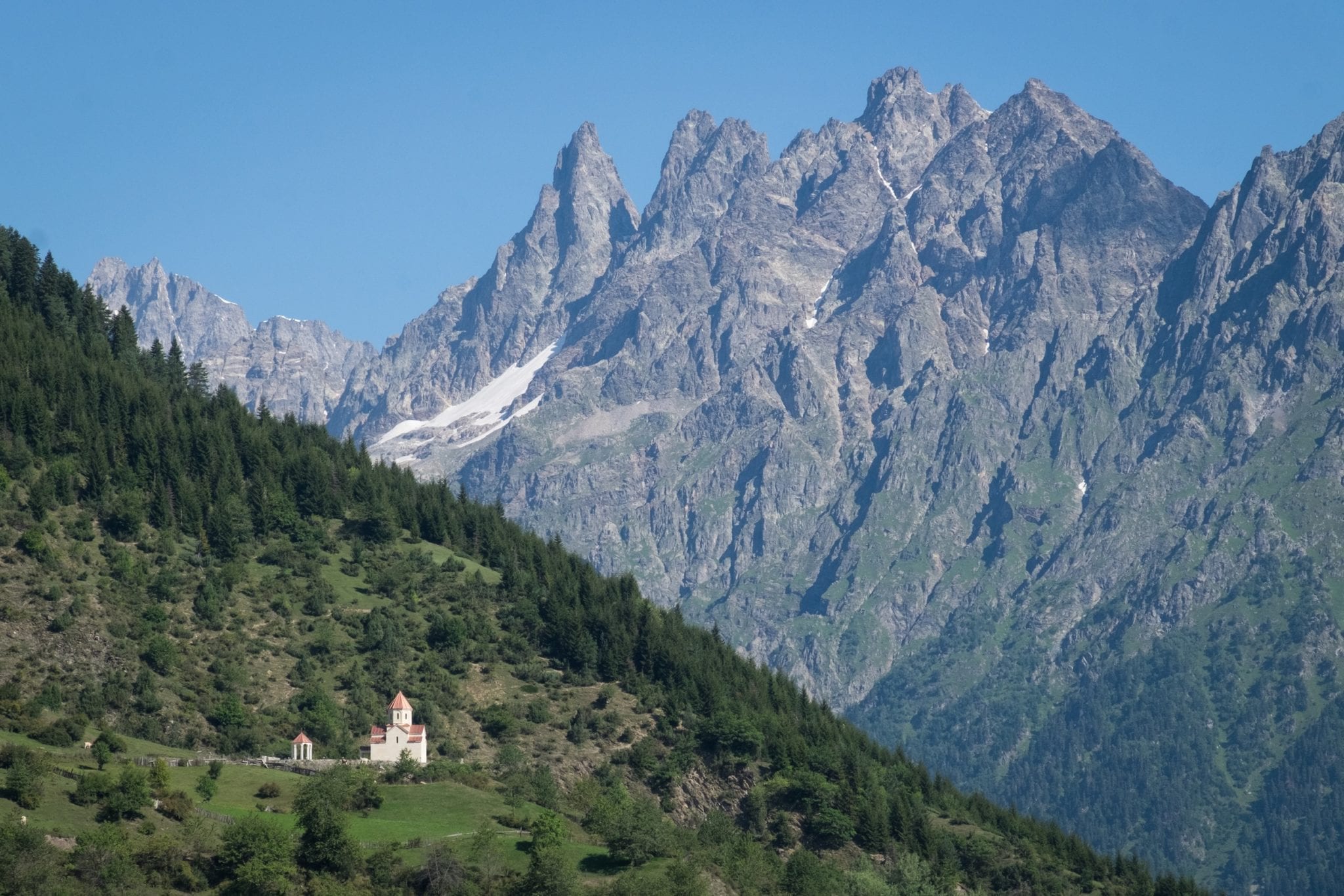 In the foreground, a green hill with a tiny church on top; in the background, a jagged gray mountain against a blue sky.