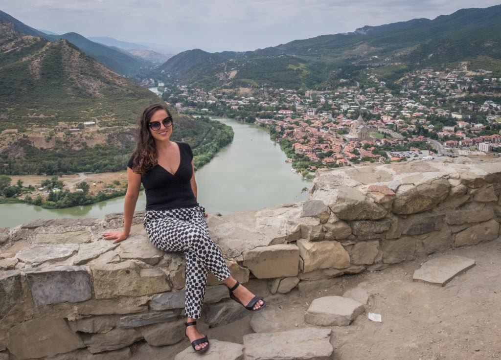Kate sits on a stone wall in front of a view of the river and the city of Mtsketa, Georgia, all green and brown in the distance.