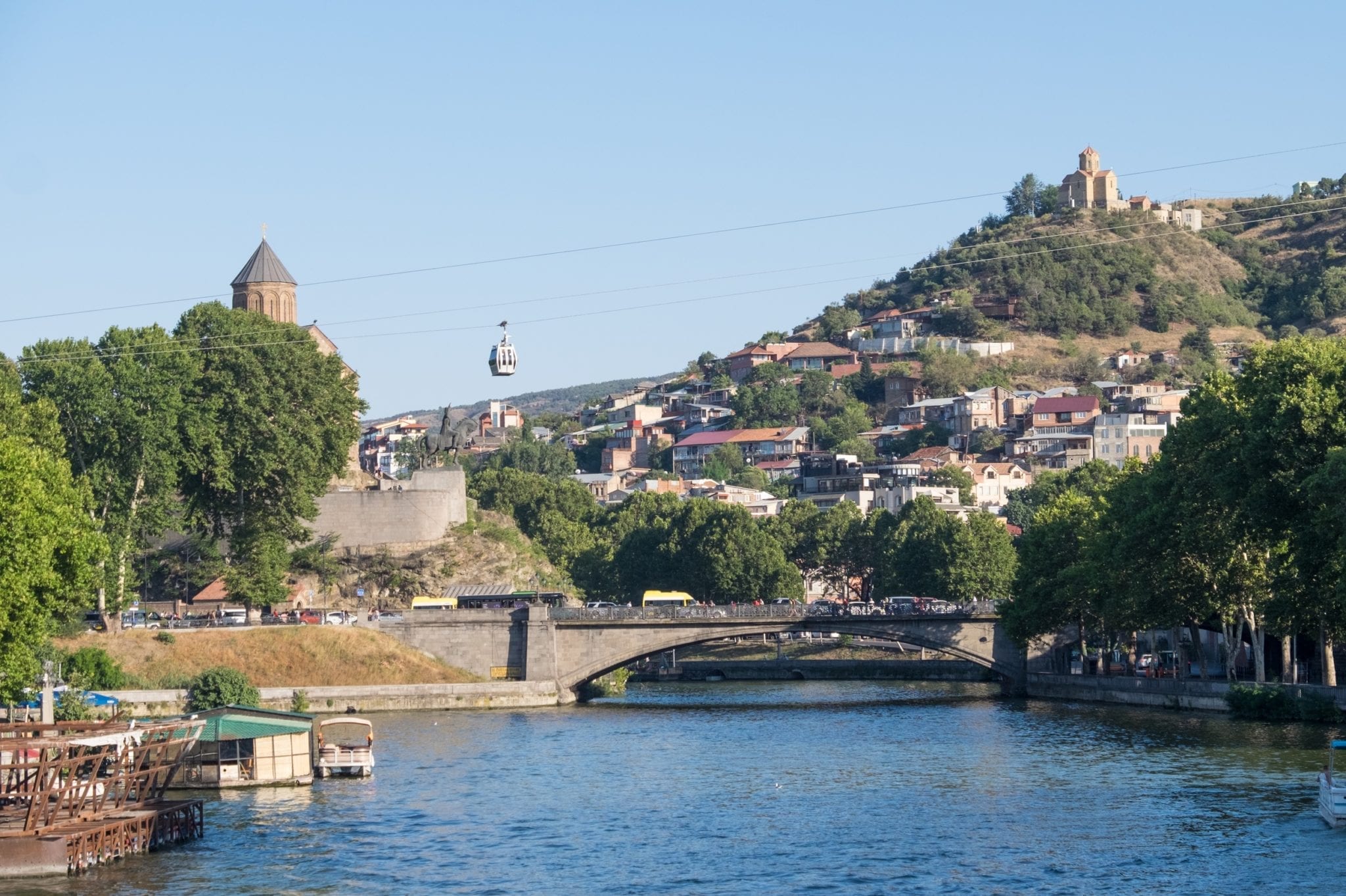 A shot over the river in Tbilisi, bridges crossing it. On the right is a monastery perched on a hill and you see a cable car traveling a wire in the air.