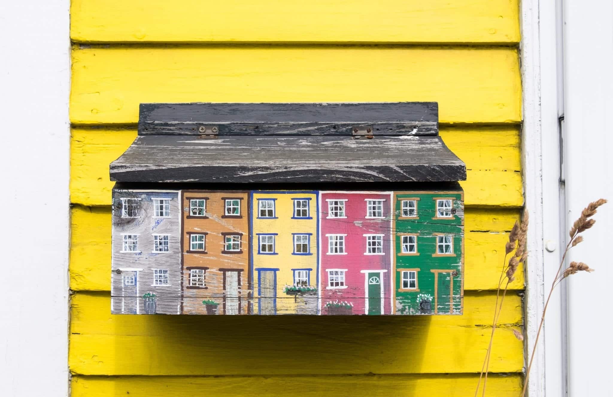 A mailbox shaped and painted like the houses of Jelly Bean Row, attached to a yellow house.