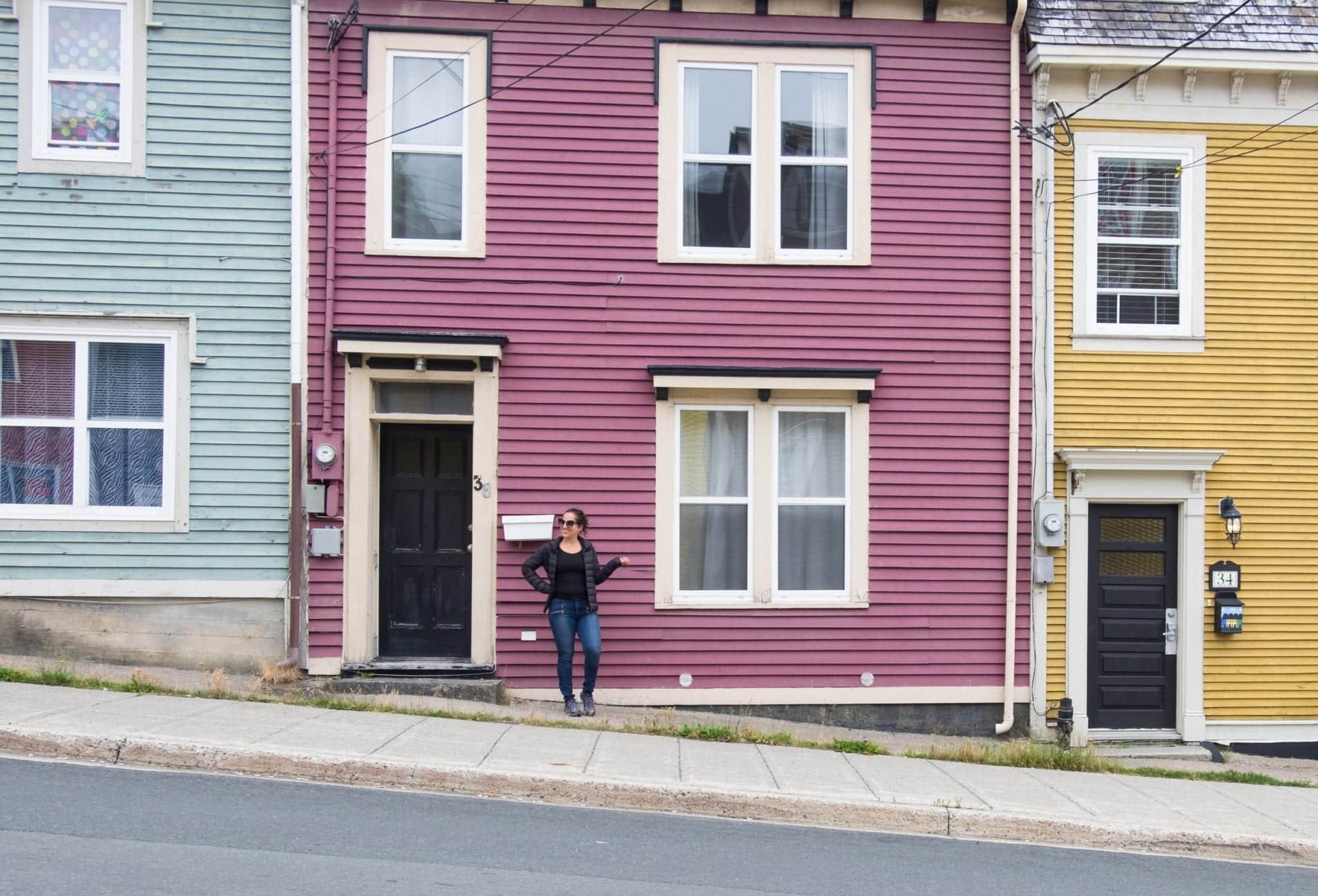 Kate stands in front of a mauve house with a pale blue house on the left and a mustard yellow house on the right.