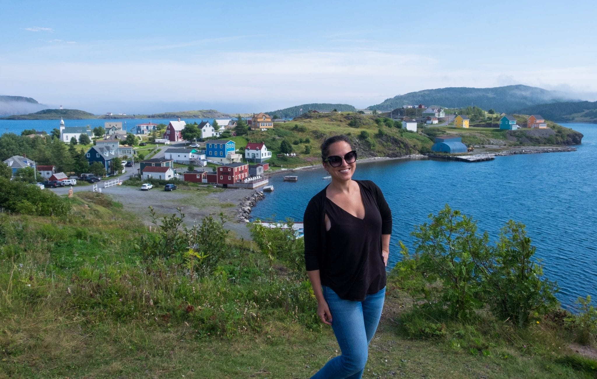 Kate stands in front of the a colorful village on a grassy peninsula surrounded by a bright blue bay.