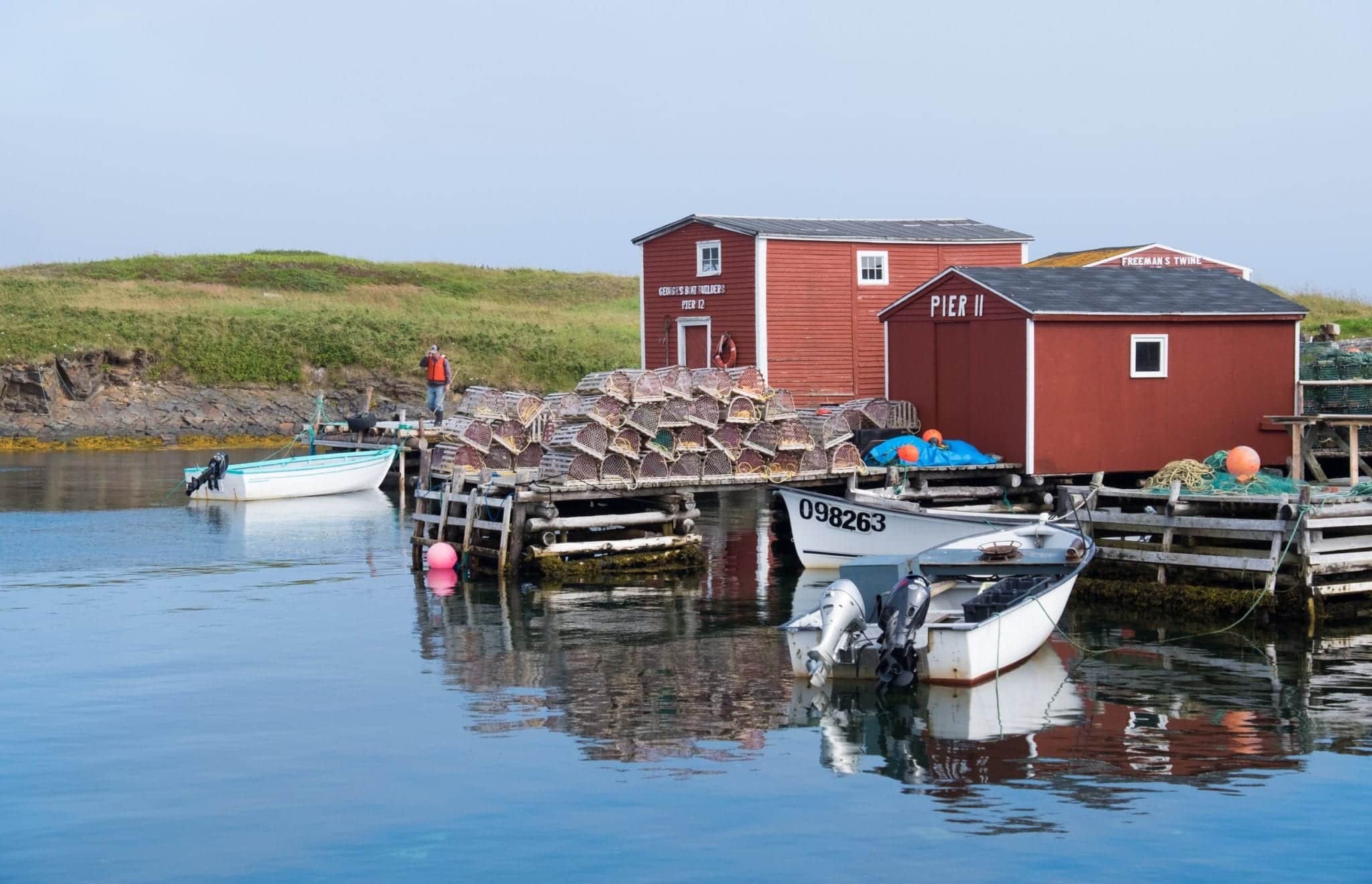 Red fishing cottages perched on the glassy blue bay next to lobster traps in Newfoundland.