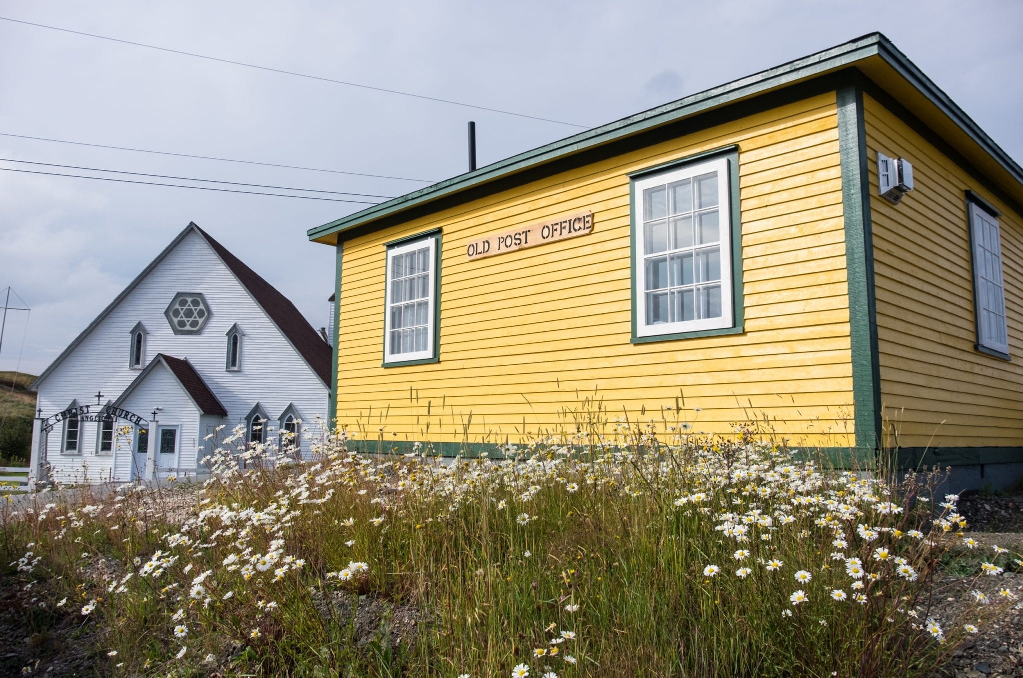 A bright yellow cottage reading "Old Post Office" in front of a field of white wildflowers, next to a white church.