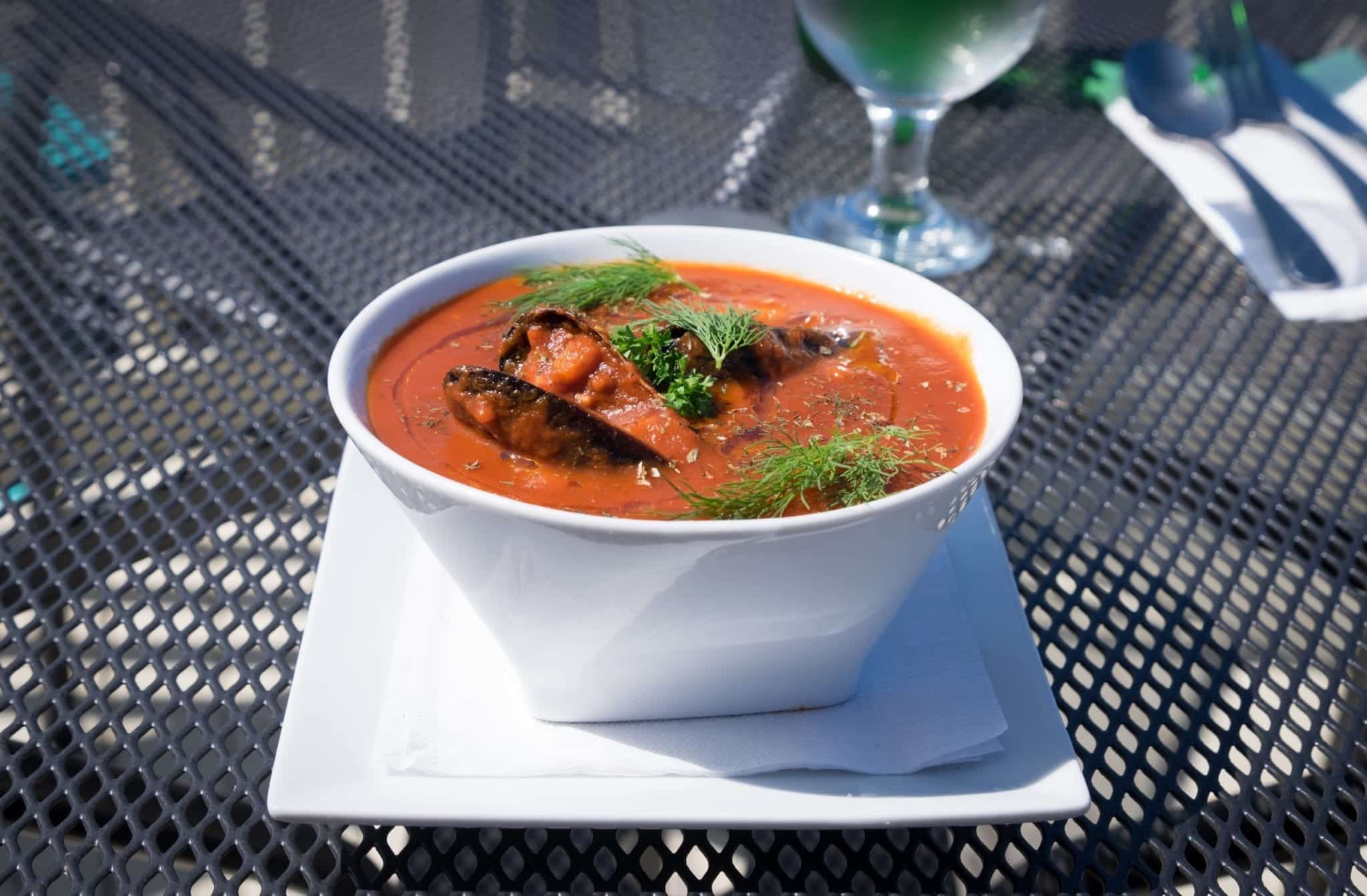 A white bowl of bright red seafood chowder -- it looks like tomato sauce with mussel shells and springs of dill poking out.