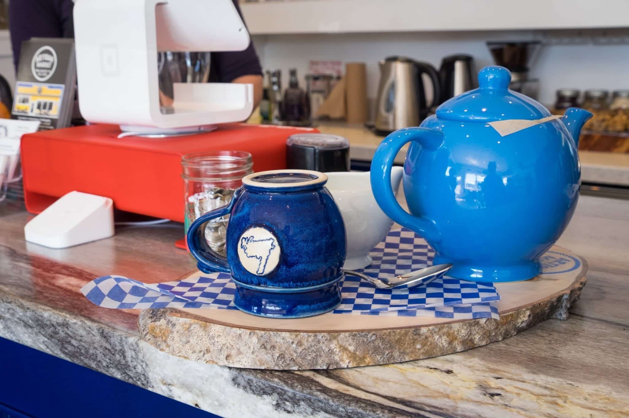 A bright blue teapot and dark blue teacup with the outline of Newfoundland on it, served on a slab of wood.