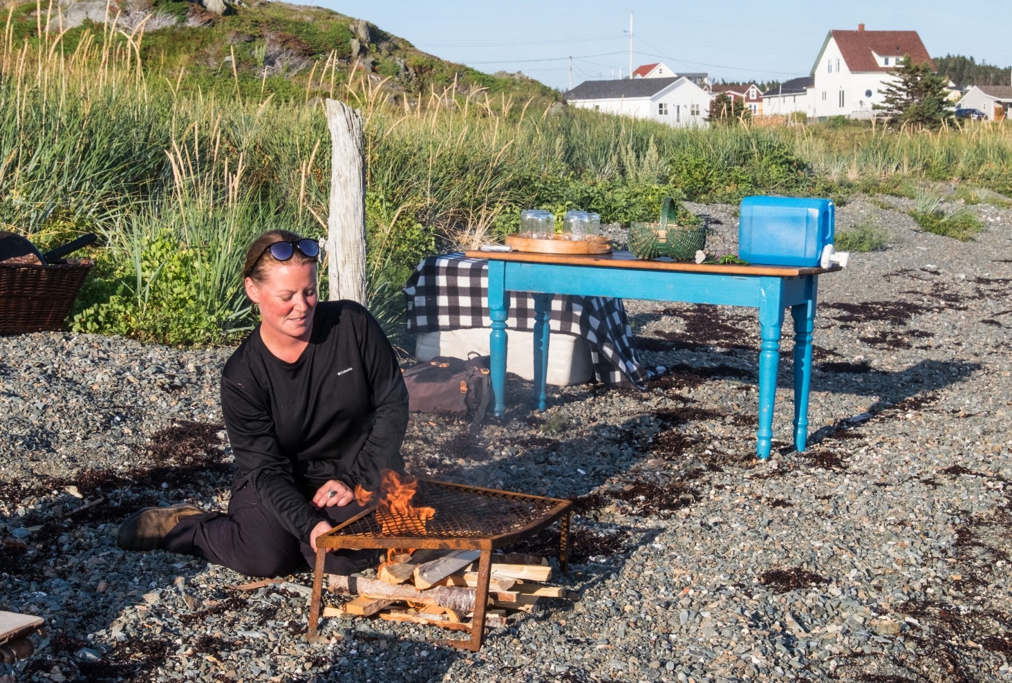 Chef Crystal tends a campfire on the beach, with cottages in the background.