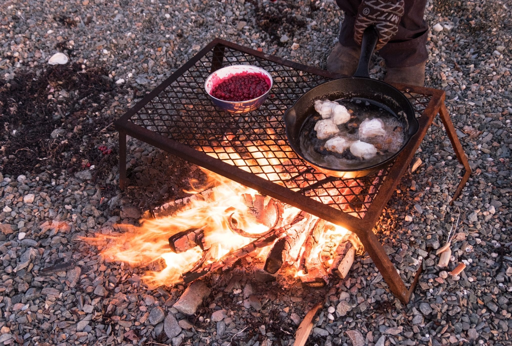 A campfire with a grill on top: cod tongues cooking in a skillet on one side, berries cooking in a dish on the other.