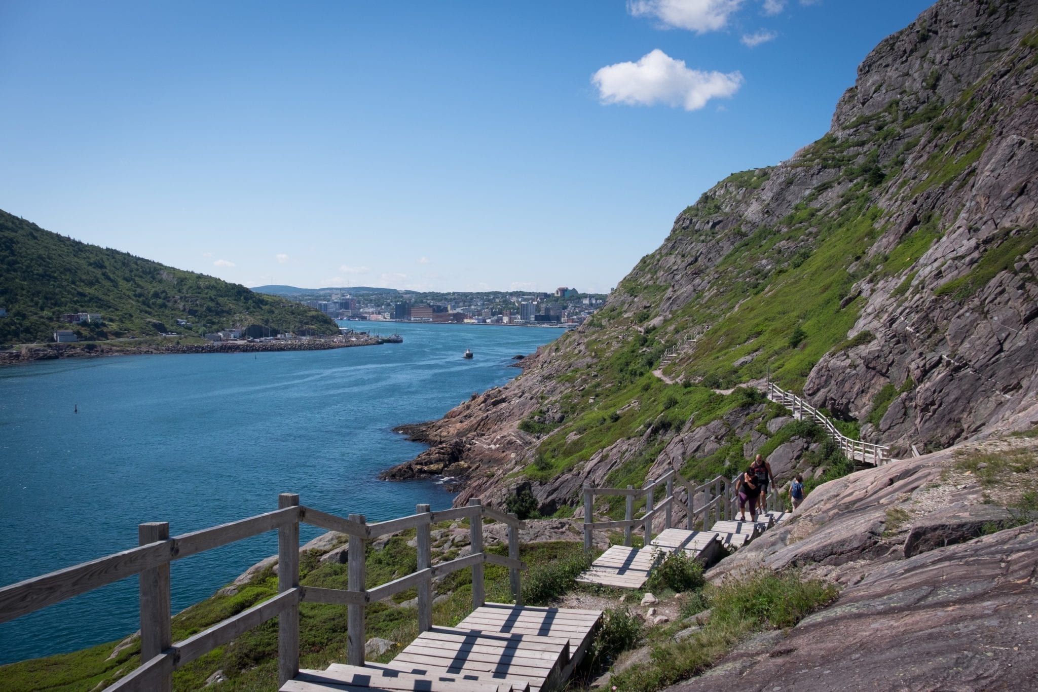 A hiking trail with wooden stairs leading up rocky cliffs in front of the bright blue bay in St. John's, Newfoundland.