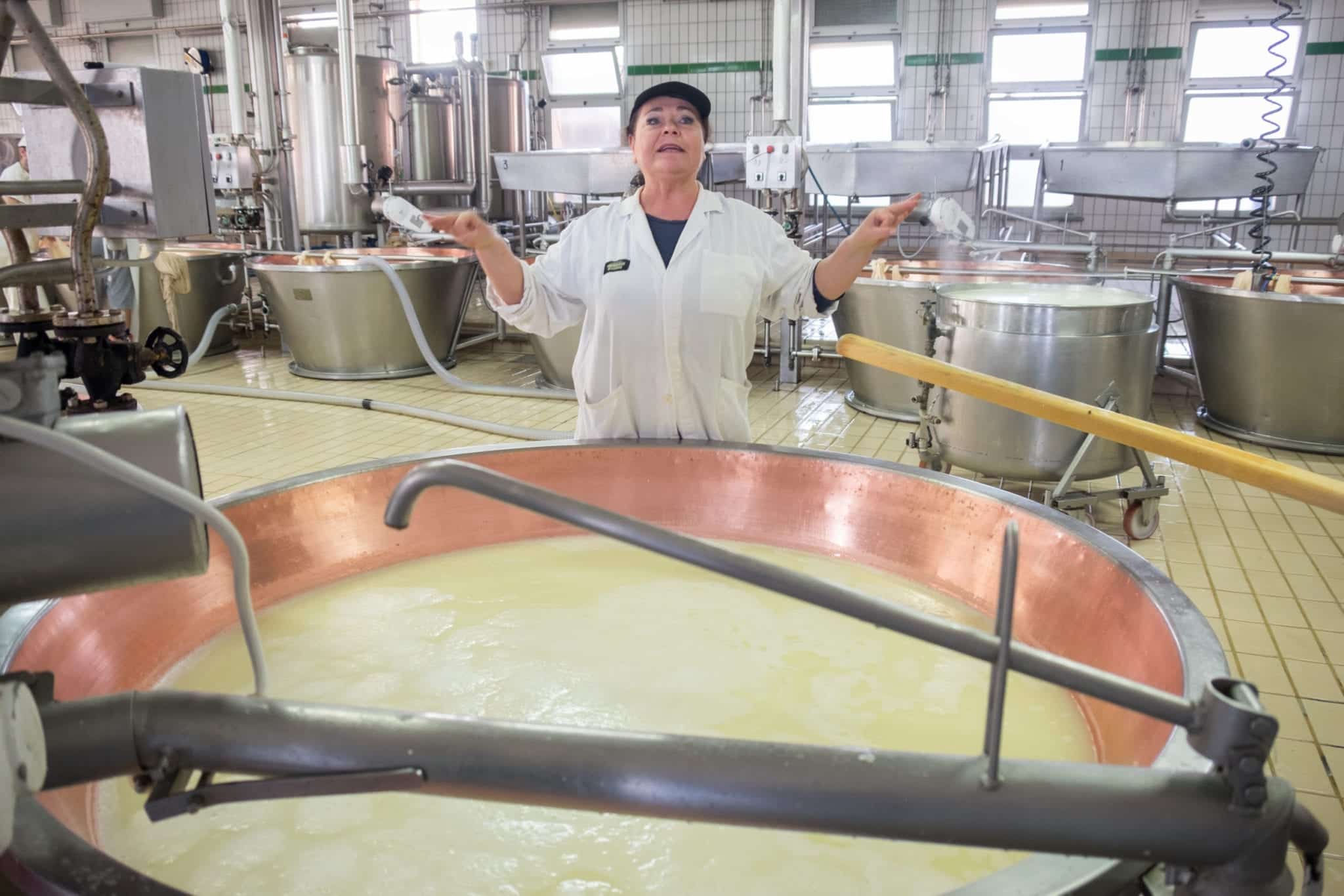A woman in a smock stands in front of a liquid vat, spreading her arms and talking about cheese.