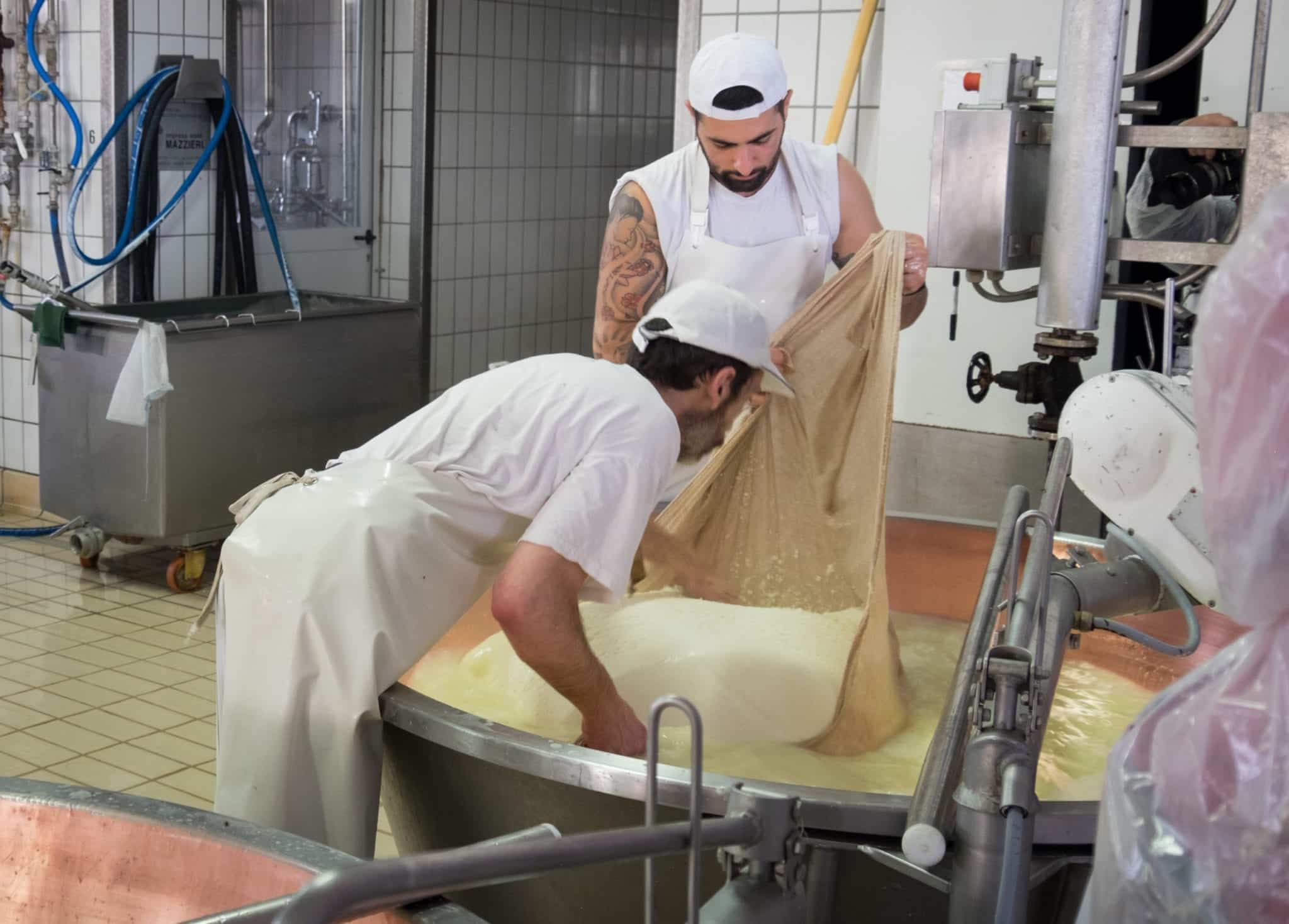 Two men, one very handsome and covered with tattoos, hold a cloth with a block of cheese on top in the vat.