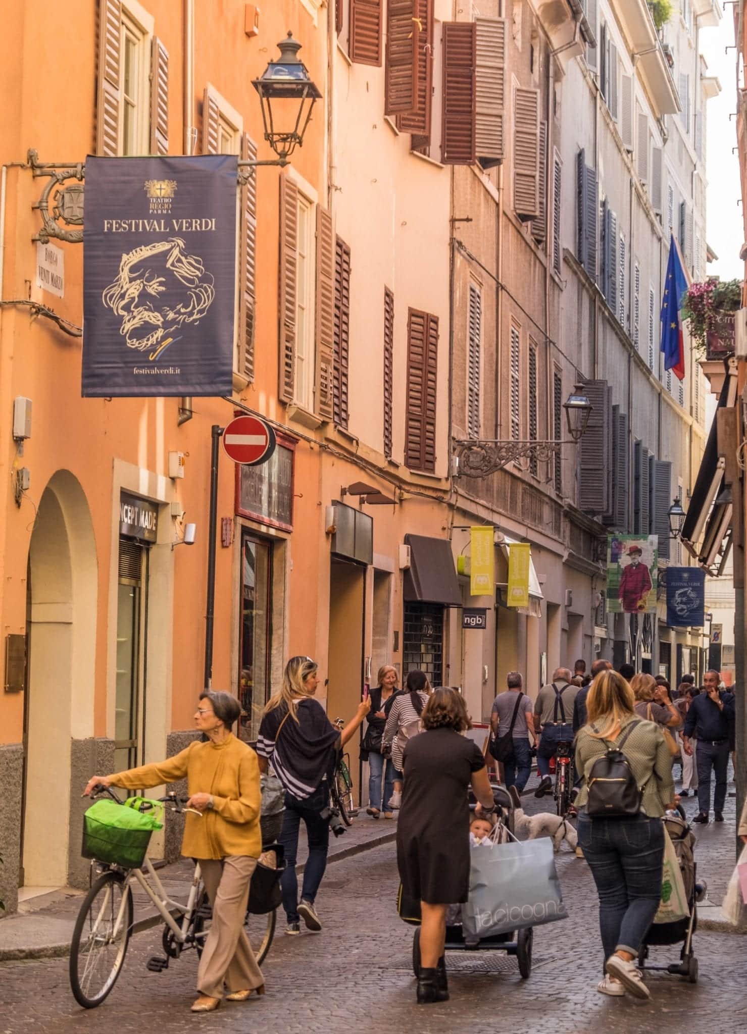 People walking down a narrow street, some walking bicycles. The buildings are warm shades of orange and pink.