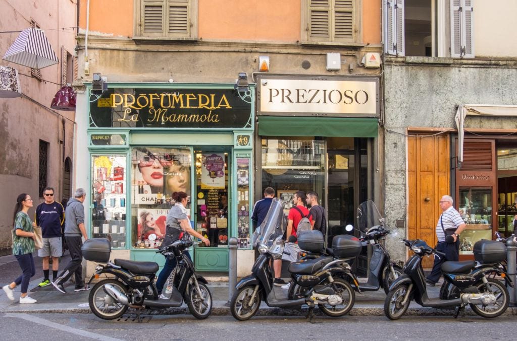 Old-fashioned stores painted green side by side in Parma, Italy, with motorbikes parked in front and people walking by.