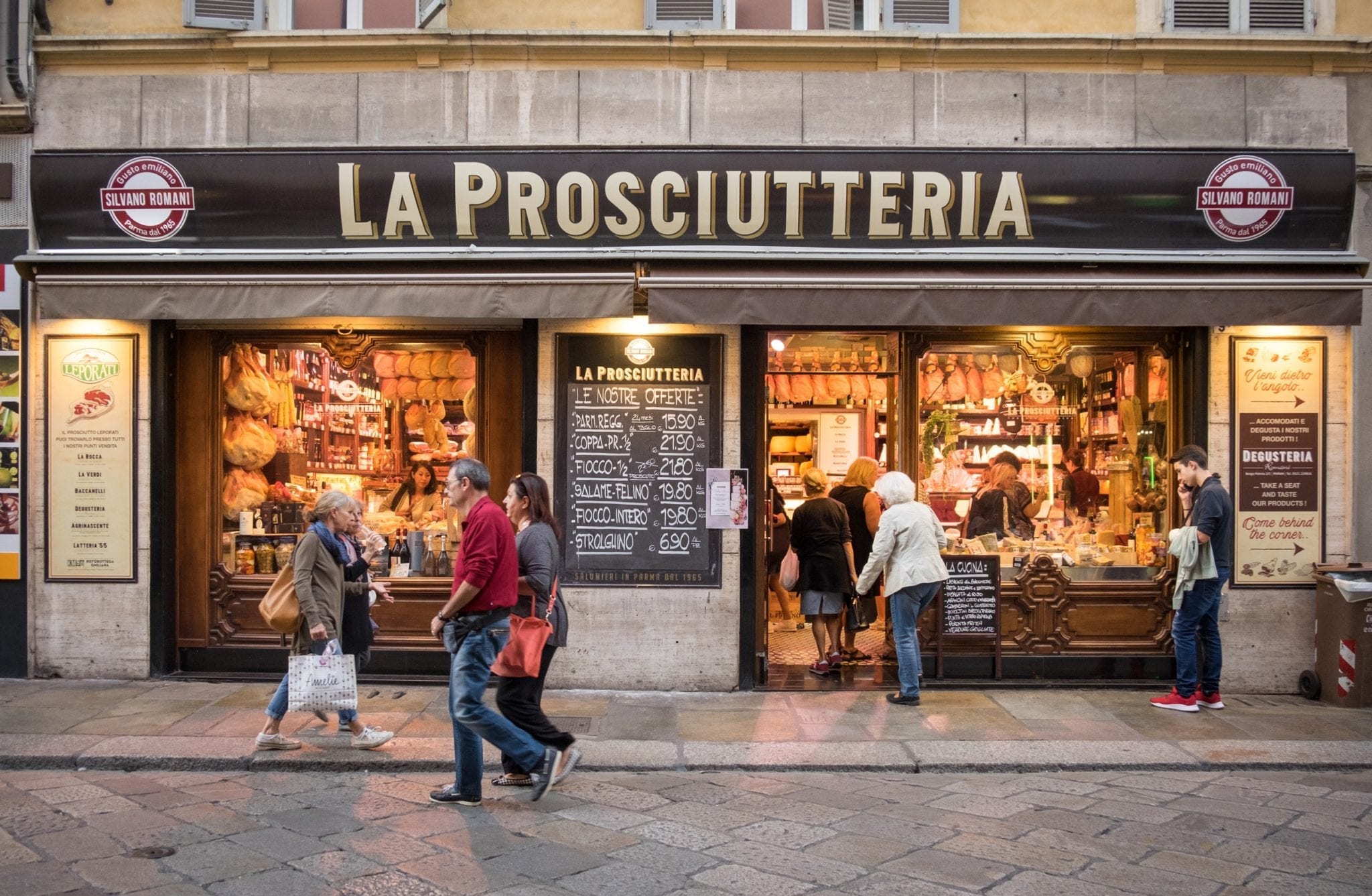 A butcher shop reading "La Prosciutteria" with people walking in front of it in Parma, Italy.