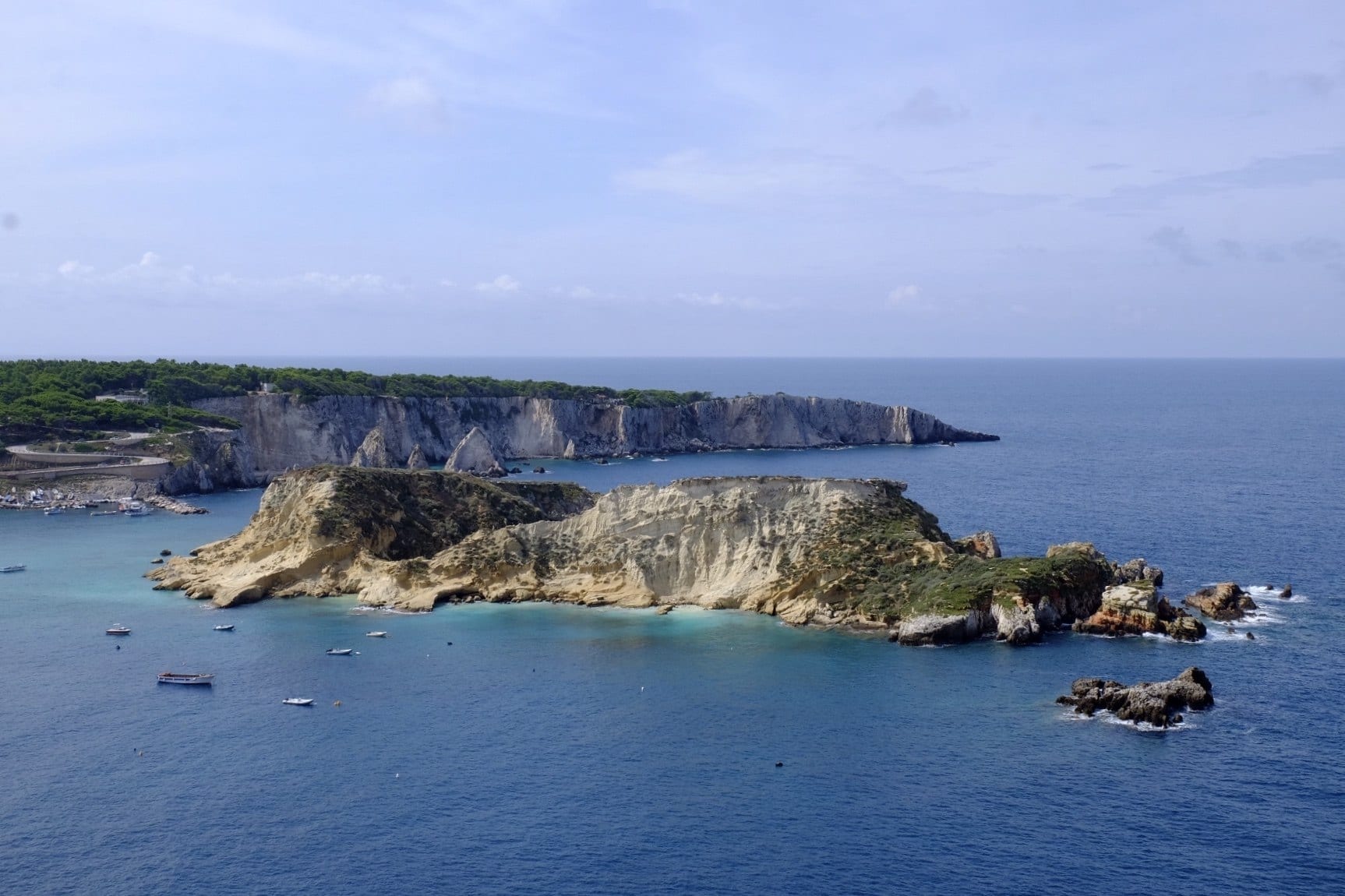 A view over two of the Tremiti Islands -- a rocky, uninhabited island, covered with sand and grass, rising out of the bright blue sea. Rowboats in the water in front of the island.
