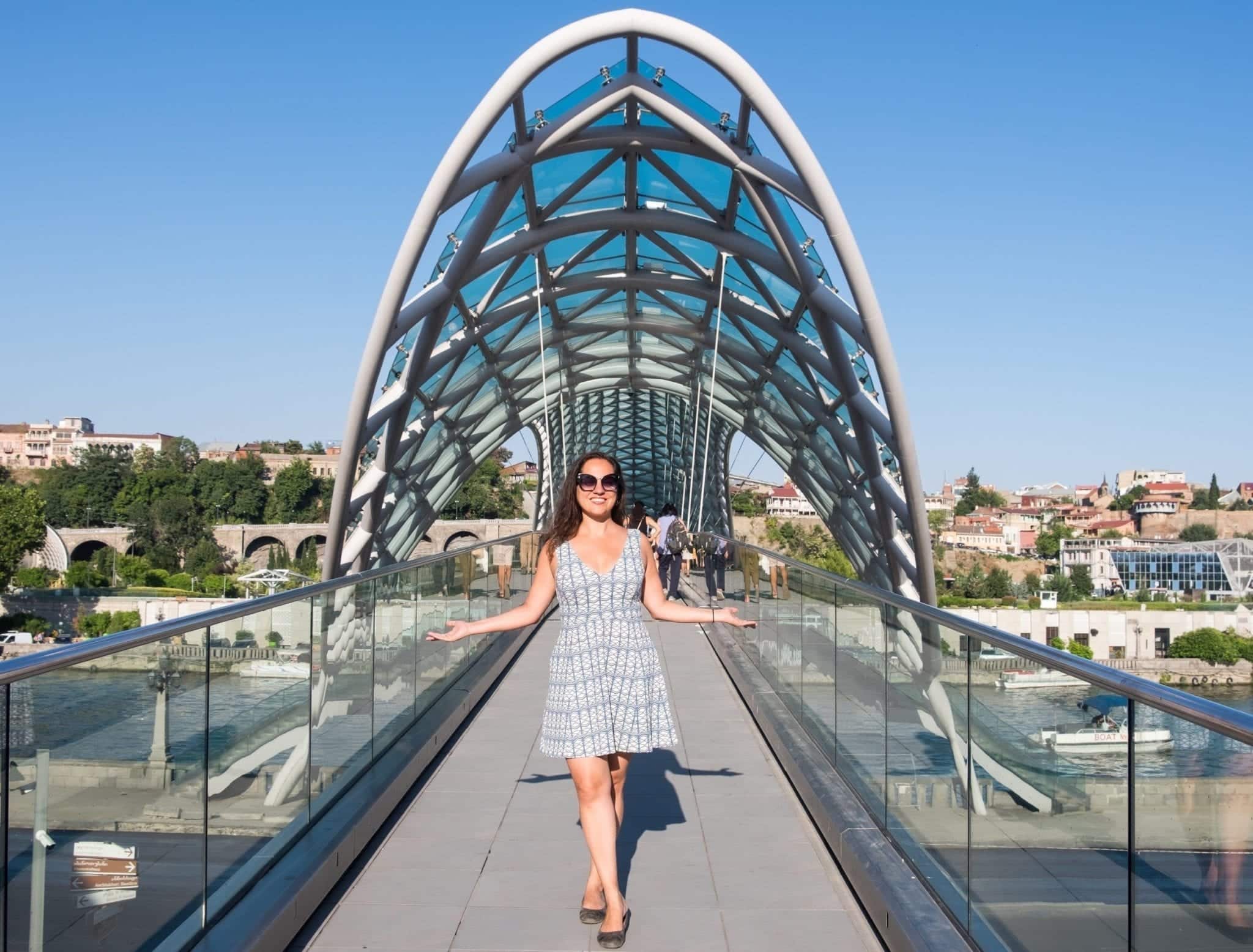 Kate stands in front of the Peace Bridge in Tbilisi, Georgia, which is modern and a grid-like oblong shape interspersed with green-blue panels of glass. Kate wears a white dress with a white and green and blue geometric pattern that looks similar to the bridge.