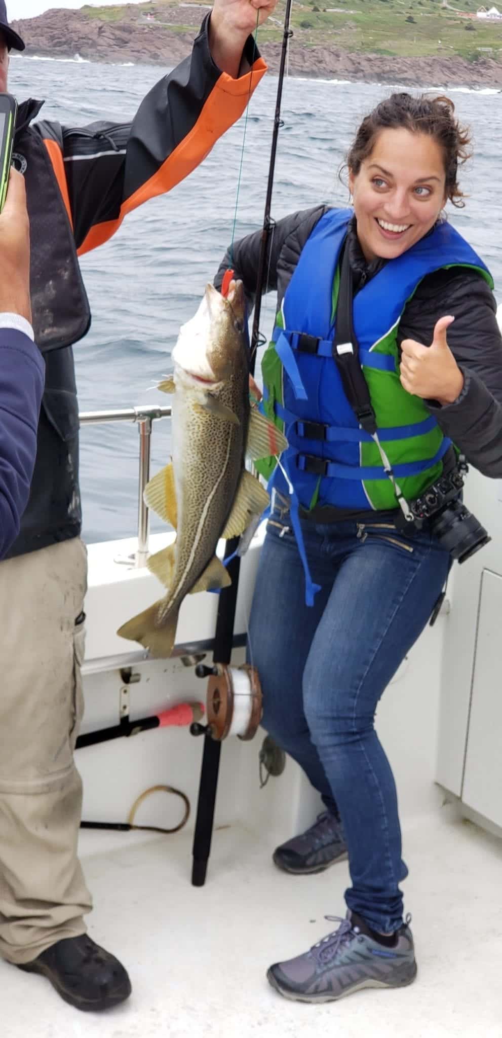 Kate holds up a fish roughly from her chest to her knees and gives a thumbs up.