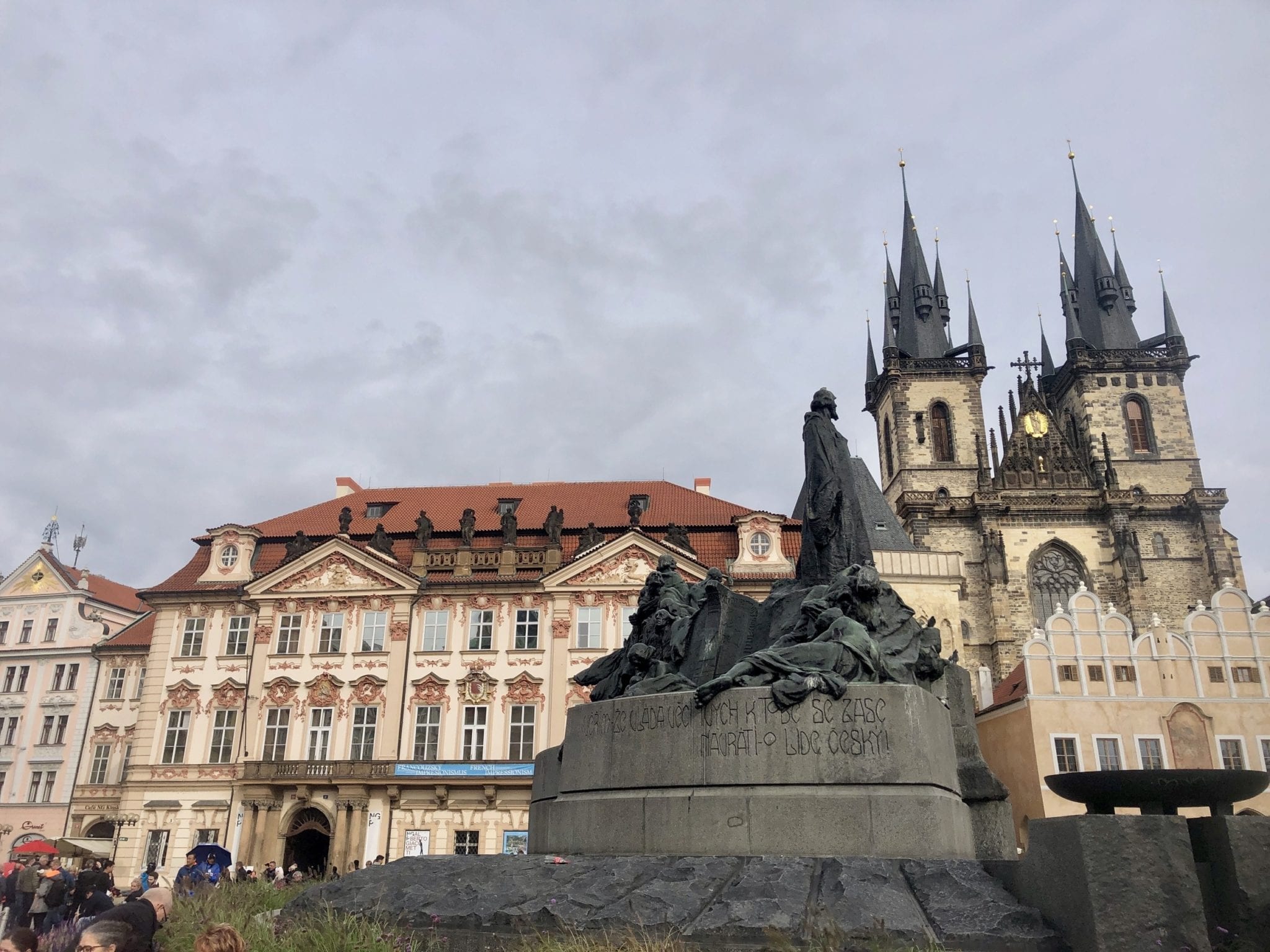 A scene from Old Town Square in Prague: a statue of soldiers in front of a cream-colored building with an orange roof and two church towers. Shot from below because THIS SQUARE IS FULL OF THOUSANDS OF TOURISTS.