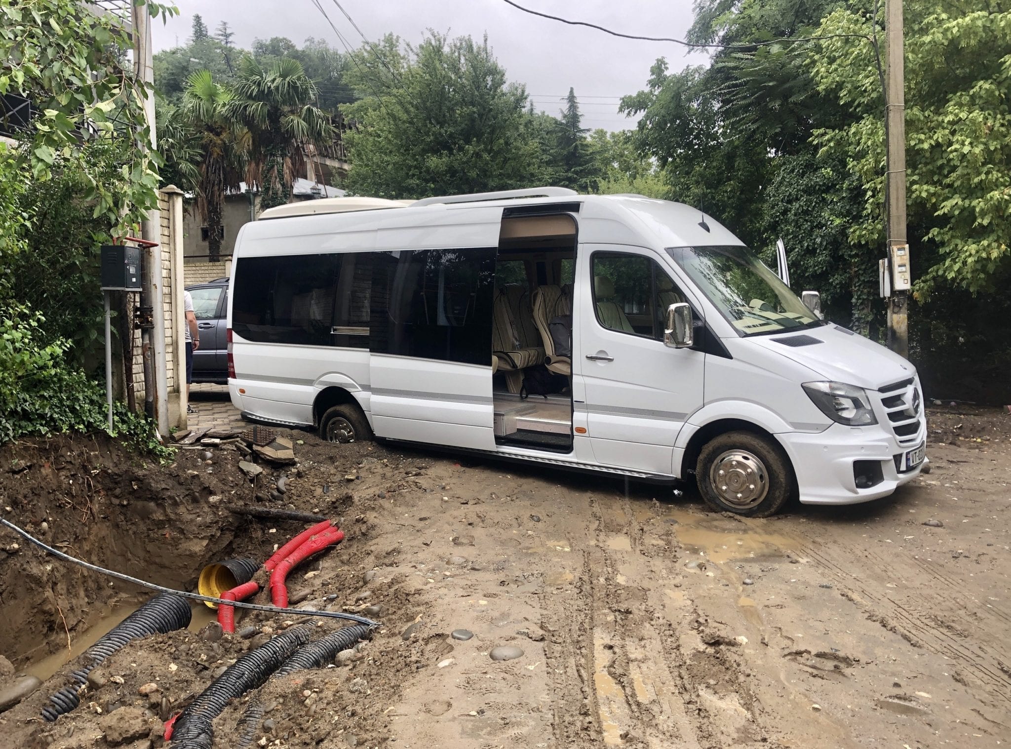 A white van on a torn-up muddy road, its back wheel sunken deeply into the mud.